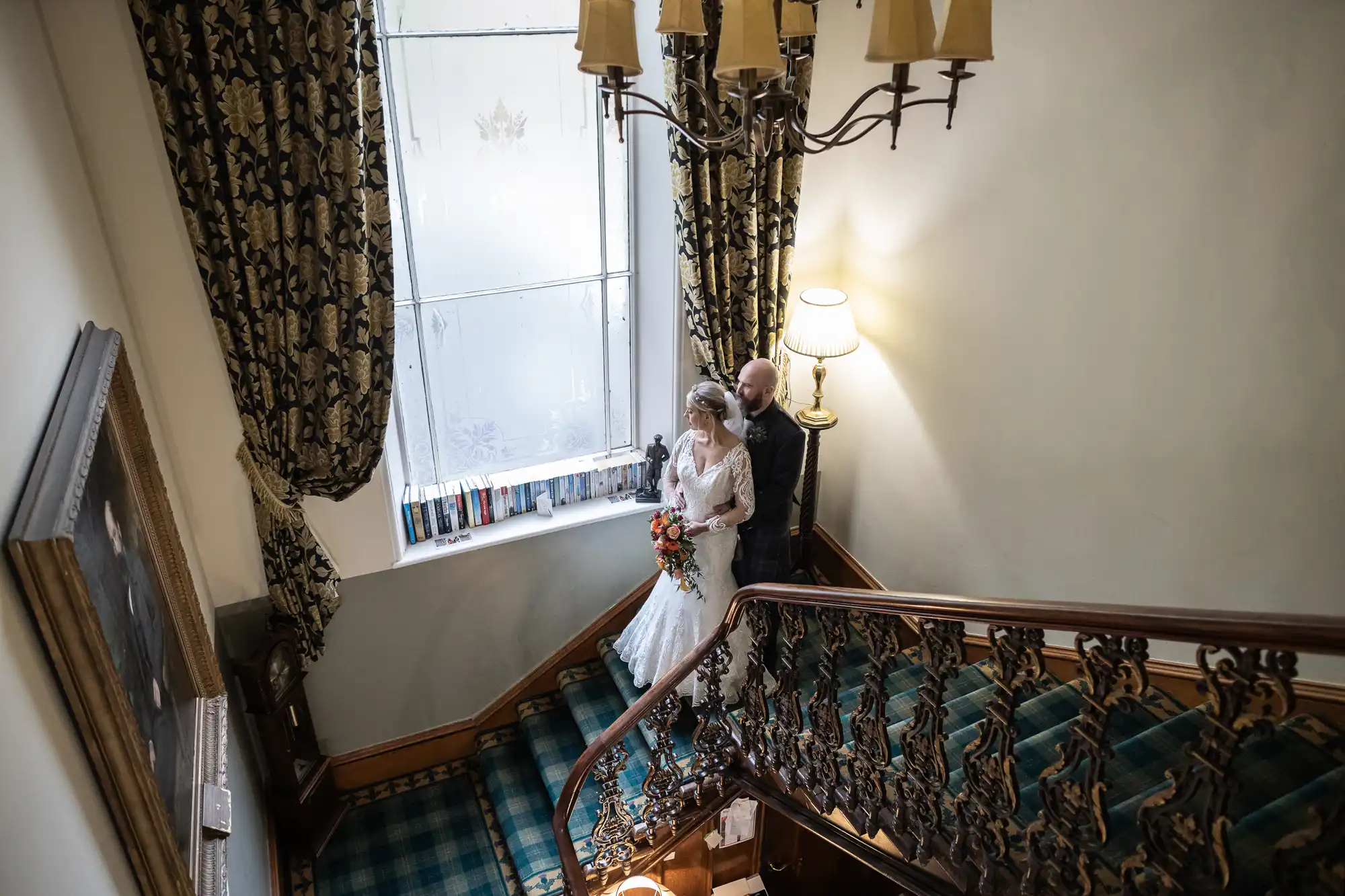 A bride in a white dress and veil holds a bouquet while walking up a staircase with a man in a suit. The staircase is carpeted with green tartan. Ornate curtains frame a large frosted window, and a lamp glows beside them. A chandelier hangs overhead.