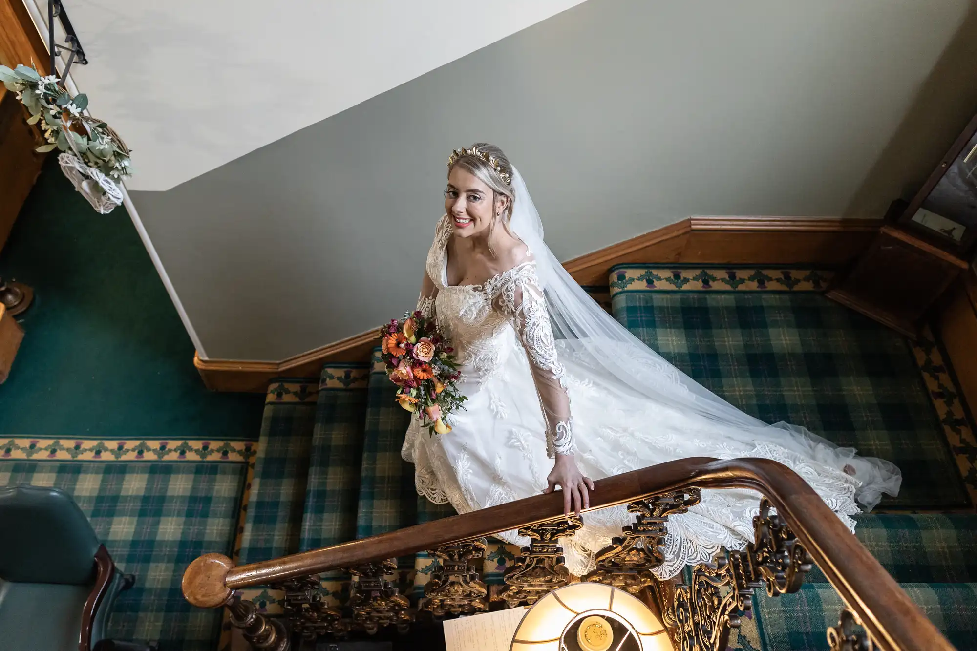 A bride in a white lace gown and veil holds a bouquet of colourful flowers while smiling up at the camera. She stands on a carpeted staircase with ornate wooden bannister in a warmly lit interior, with green and blue tartan carpet underfoot.