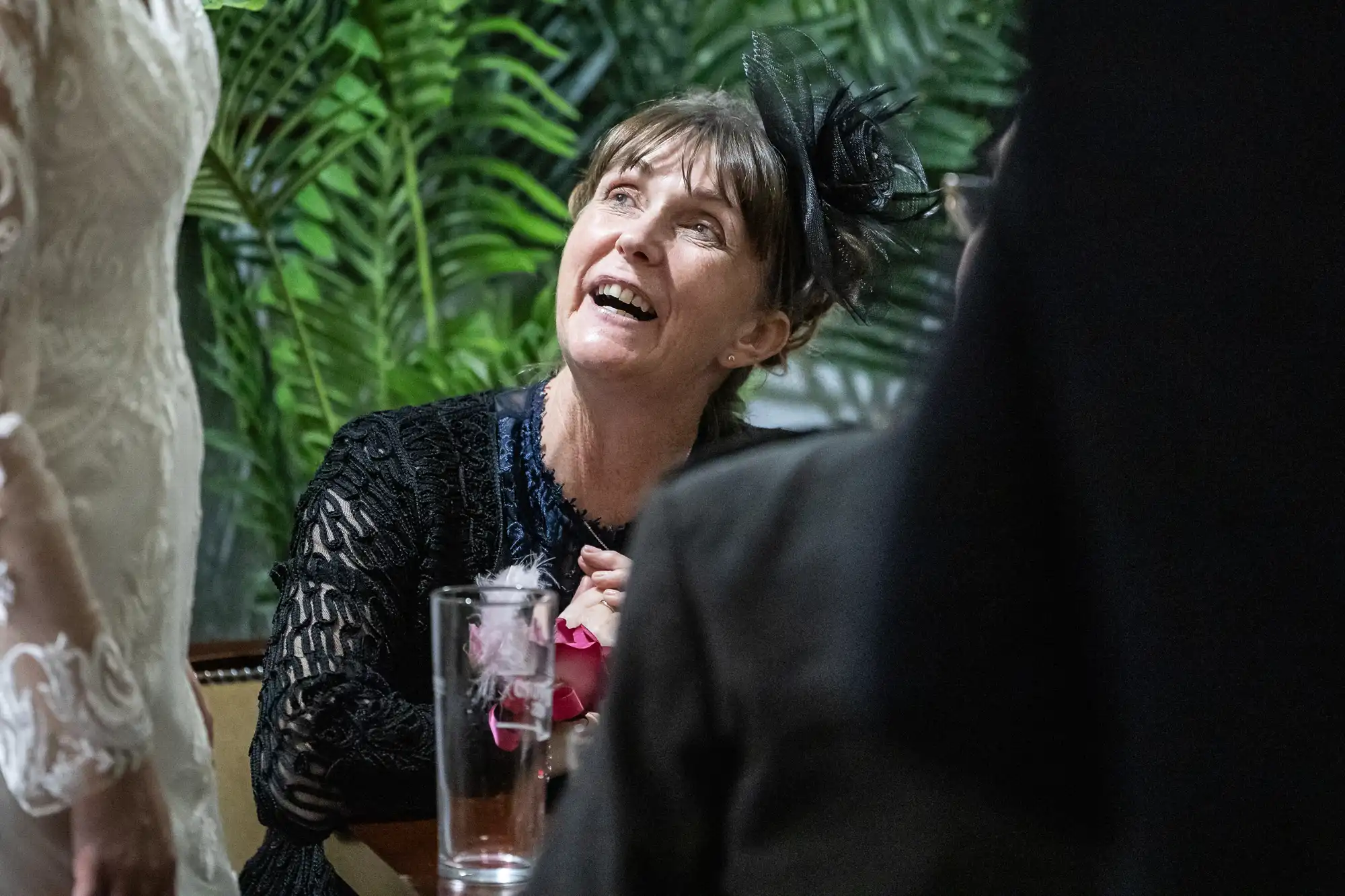 A woman in a black dress and fascinator sits at a table, smiling and looking up. She holds a drink with a pink flower inside. Green leafy plants are in the background, and a person in a white lace sleeve stands beside her.