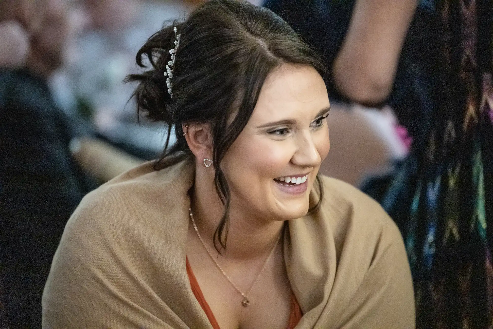 A woman with dark brown hair styled in an updo with hair accessories smiles brightly. She wears a tan shawl over a rust-coloured top and a heart pendant necklace. The background is softly blurred, suggesting an indoor social event.