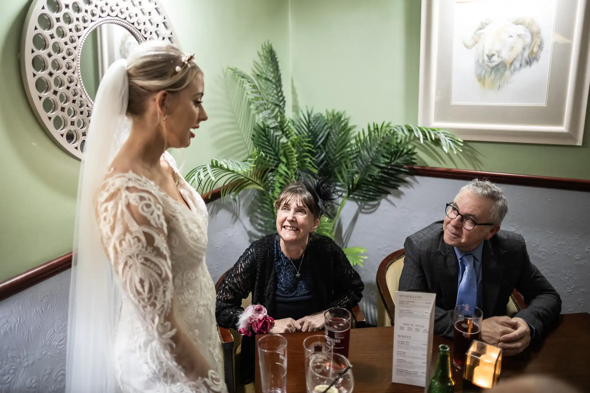 A bride in a white lace dress and veil stands smiling, talking to an older woman and man seated at a table with drinks. The woman smiles up at the bride. Behind them are a plant, a round mirror, and a framed picture of a lion.