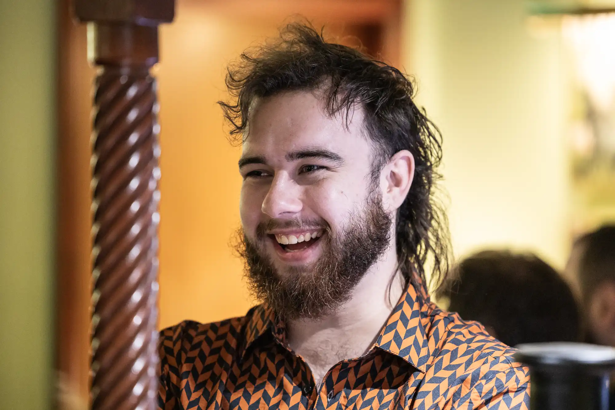 A young bearded man with a mullet hairstyle smiles broadly. He wears a patterned, orange and black button-up shirt. The background is softly lit with warm tones and slightly out of focus. A carved wooden post is visible to the left.