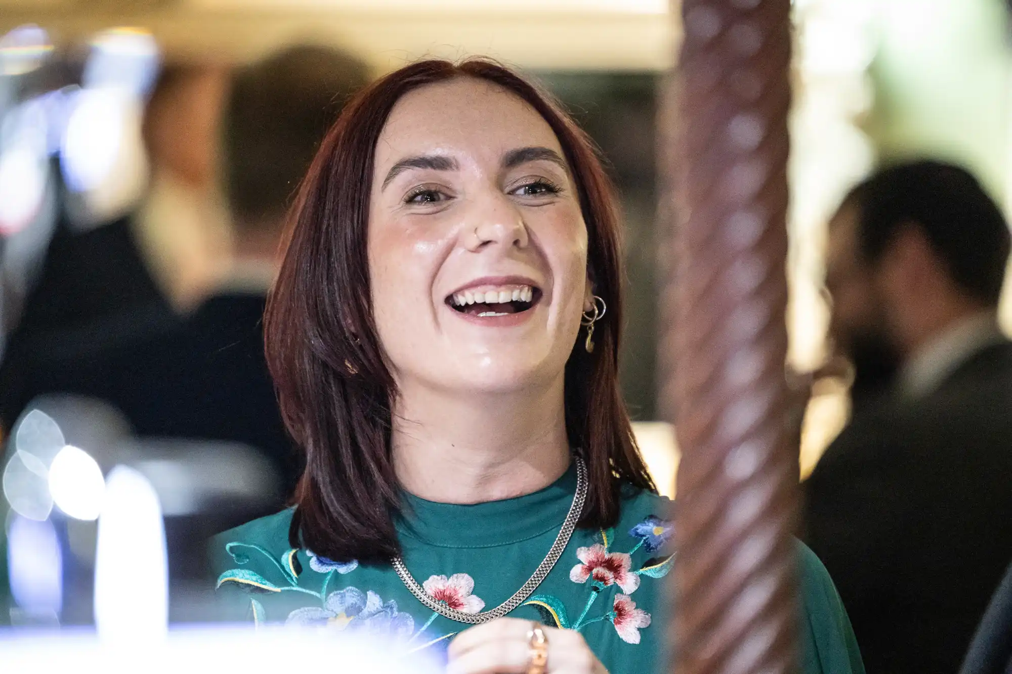 A woman with straight, shoulder-length dark hair smiles brightly. She wears a green top with colourful floral embroidery, gold necklaces, and hoop earrings. The background is softly blurred with people and lights, creating a warm, joyful atmosphere.