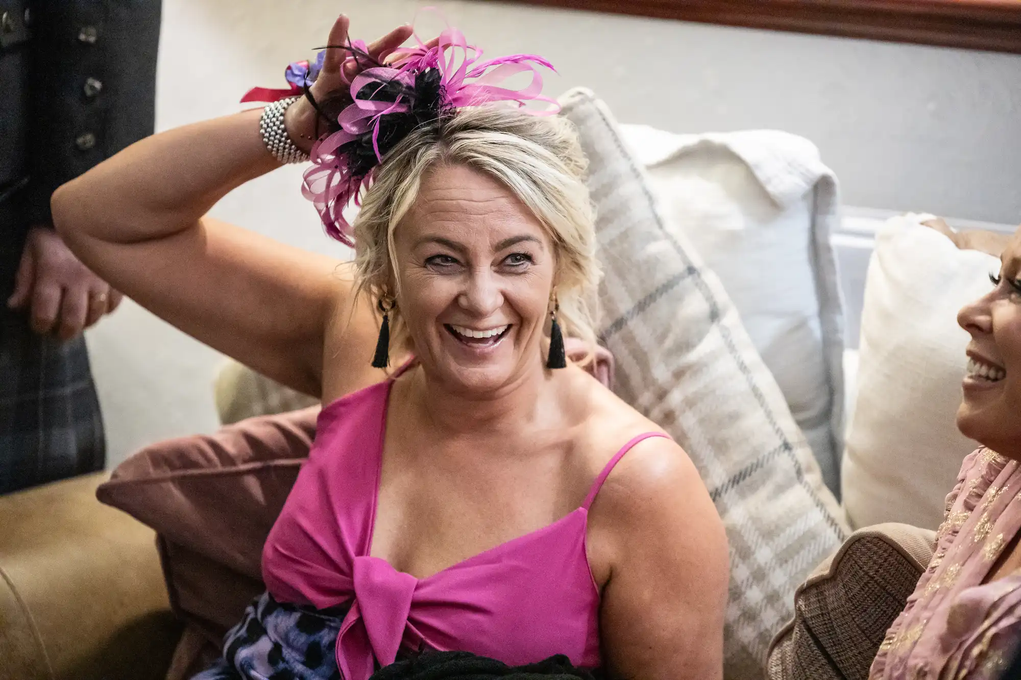 A smiling woman in a bright pink dress adjusts a pink and black fascinator on her blonde hair. She sits on a sofa with beige and checked cushions, wearing earrings and a bracelet, while another person in light clothing sits beside her, partly visible.