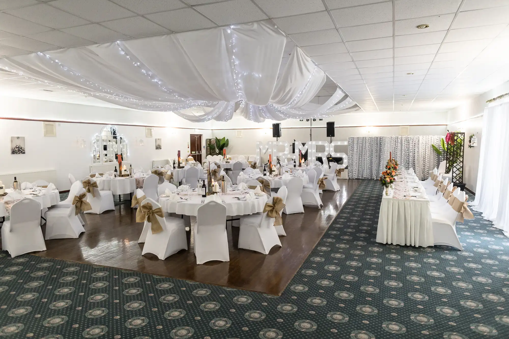 A decorated banqueting hall with round tables covered in white cloths and chairs tied with gold bows. White drapes and fairy lights hang from the ceiling. A long rectangular table is set at one side, with floral centrepieces on tables and soft lighting.