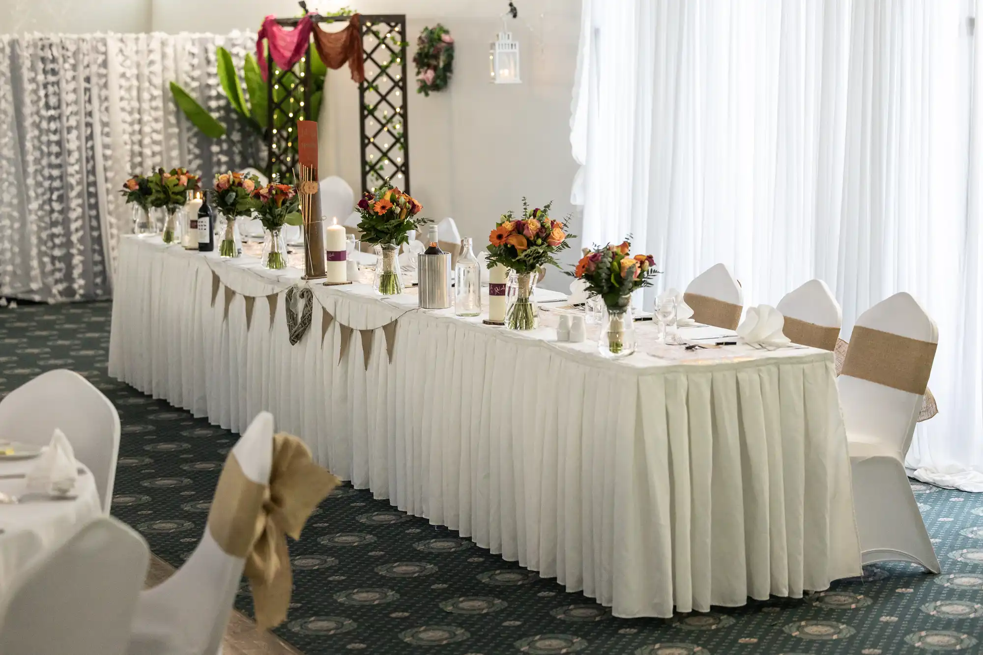 A long banquet table draped in white cloth is decorated with floral centrepieces, candles, and table settings. White chairs with beige sashes surround the table, and there are white curtains and greenery in the background, creating an elegant event space.