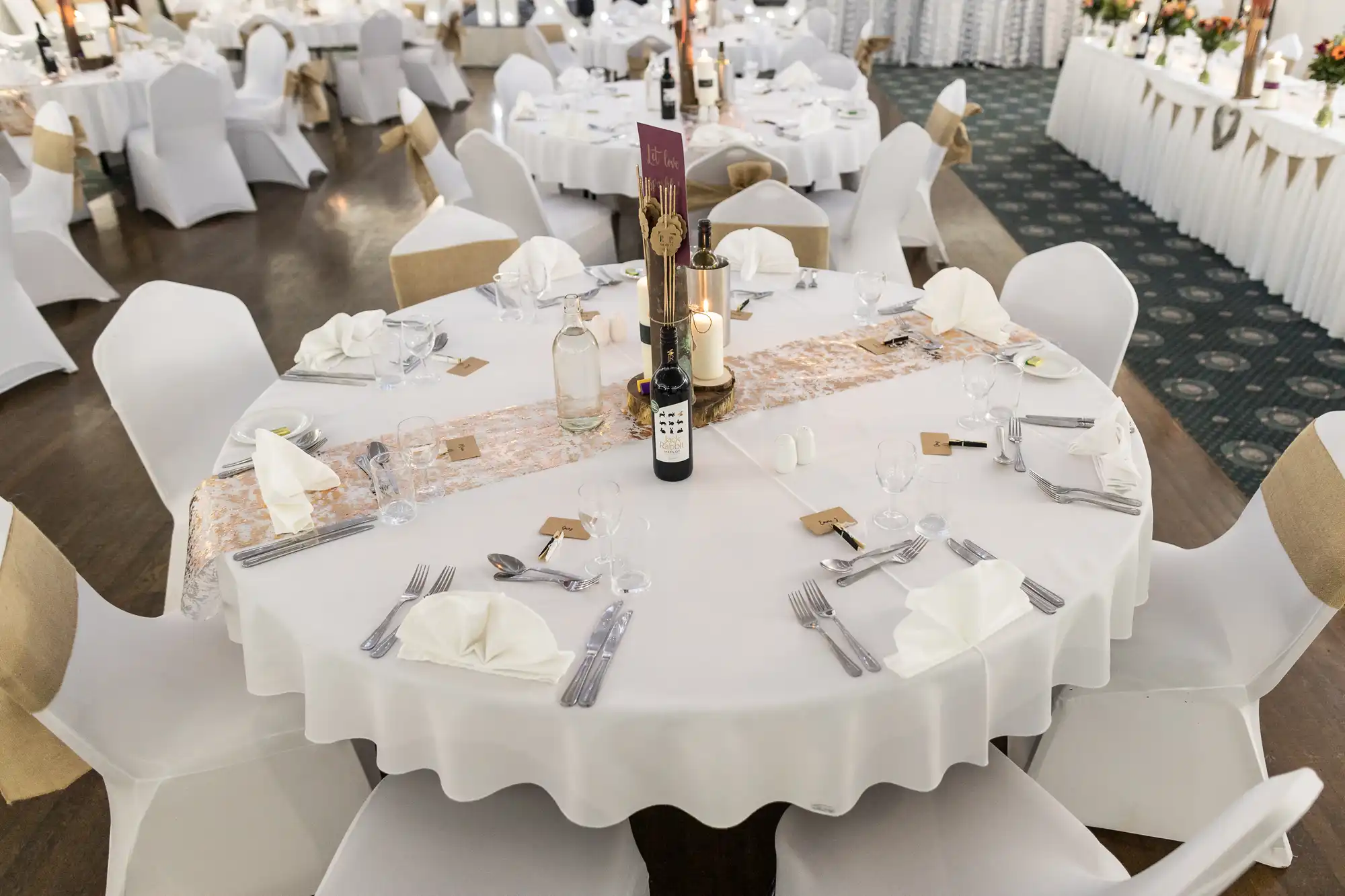 A round banquet table set for six with white cloth, folded serviettes, cutlery, wine glasses, a wine bottle, water carafe, candles, menu cards, and small place cards. Chairs are covered in white fabric with gold sashes in a decorated event space.