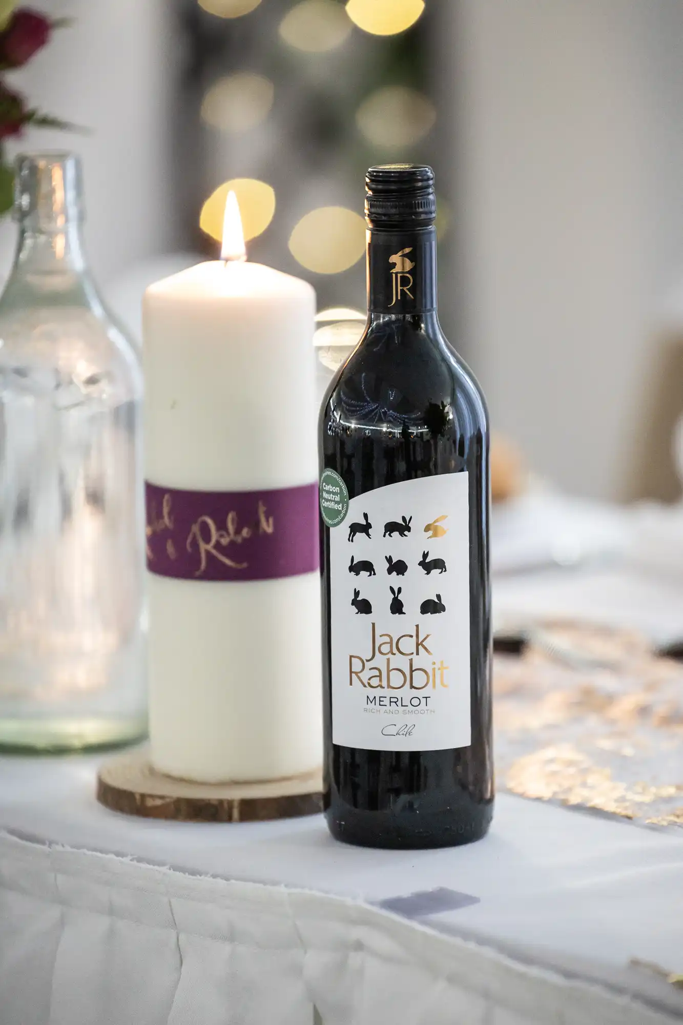 A bottle of Jack Rabbit Merlot wine stands on a white table next to a tall white candle with a purple band. The candle is lit, and there are blurred lights and glassware in the background, creating a cosy, elegant atmosphere.