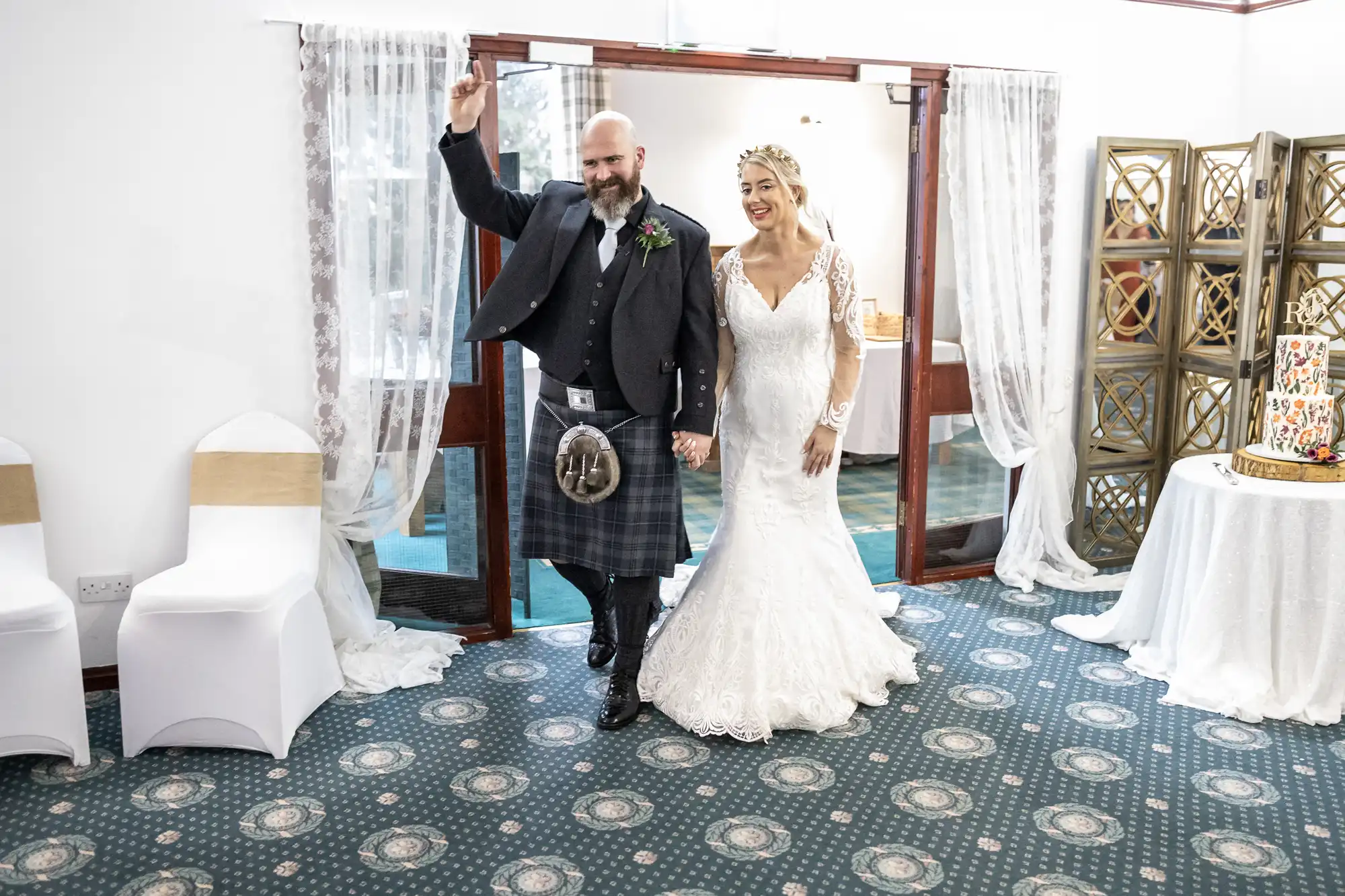 A smiling bride in a white lace gown and a groom in a black jacket and traditional Scottish kilt enter a decorated room hand-in-hand. The groom raises one arm in celebration. White-covered chairs and a table with a colourful cake are seen nearby.