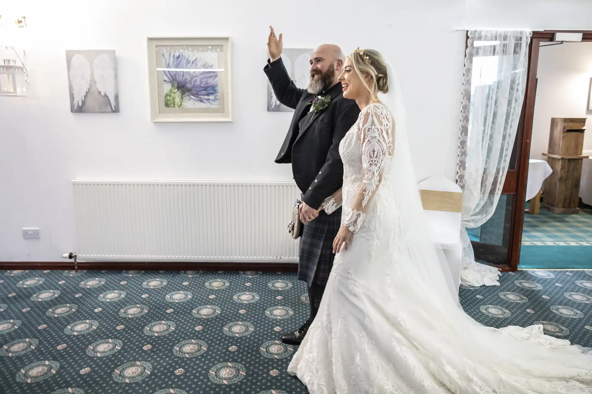 A bride in a white lace gown stands next to a bearded groom in a dark jacket and kilt. He waves with a smile. They are indoors on patterned carpet, with artwork and a radiator on the white wall behind them. Light streams from a doorway on the right.