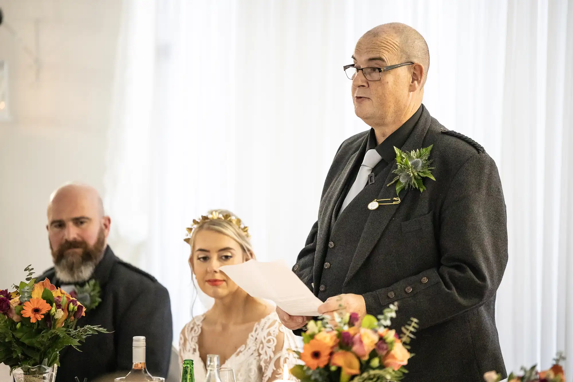 A man in a dark suit stands giving a speech, holding papers, next to a bride in a white dress and floral crown, and a bearded man. All are seated at a decorated table with flowers and drinks, in a bright room with sheer white curtains.