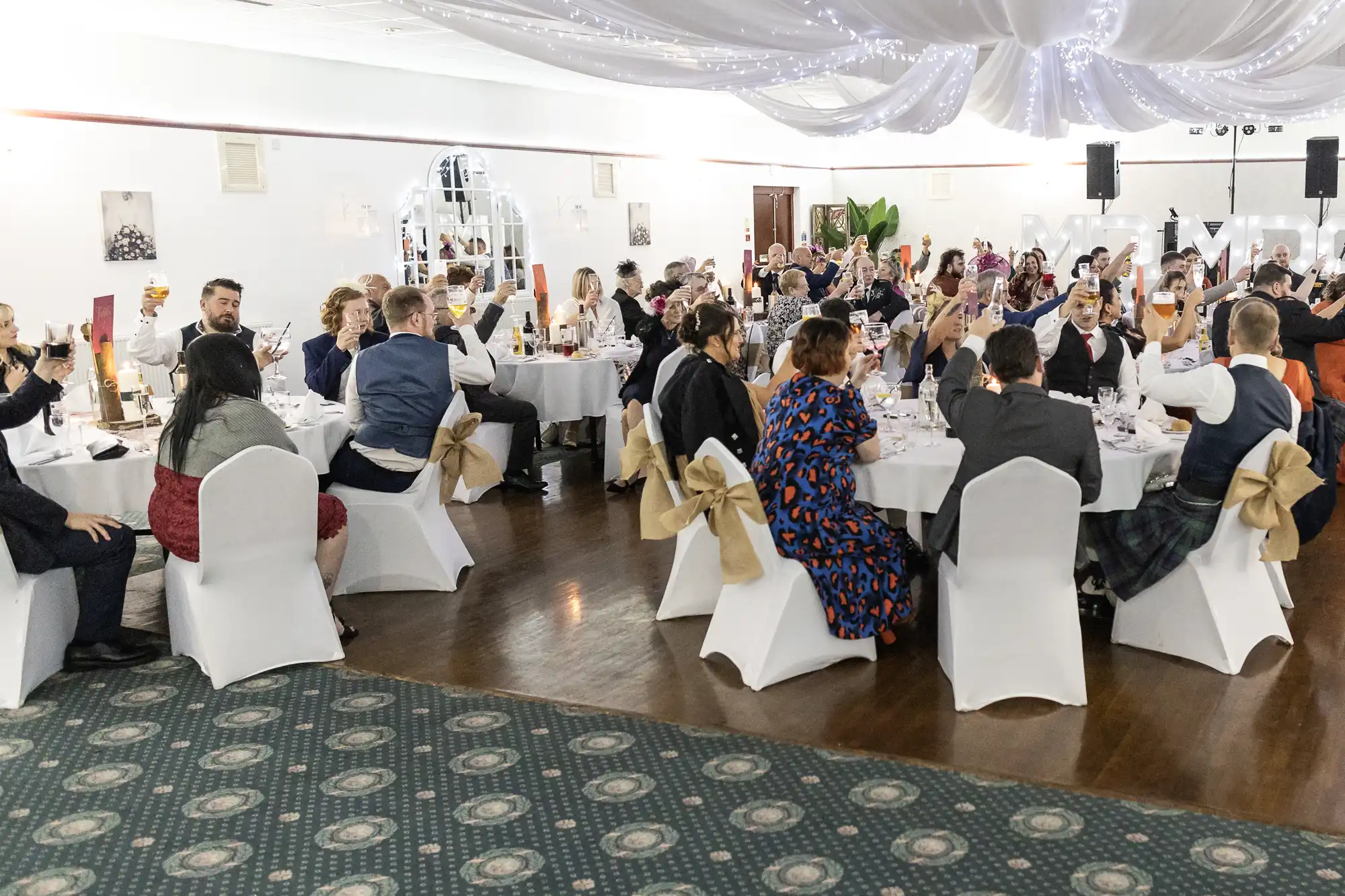A large group of people in formal attire sit at round tables in a decorated banqueting hall with white chair covers and golden bows, raising glasses in a toast under draped ceiling lights. The atmosphere appears festive and celebratory.