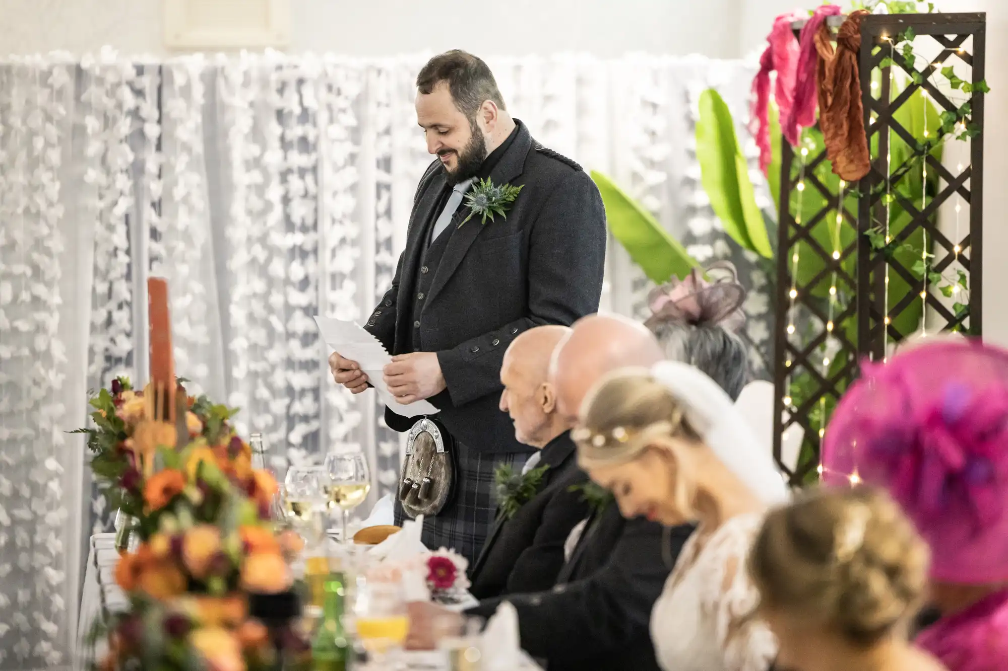 A man in a dark suit stands, reading from a paper, giving a speech at a decorated wedding table. Seated guests, including a bride in white with a veil, listen attentively. The scene is festive, with flowers and greenery on the table and in the background.