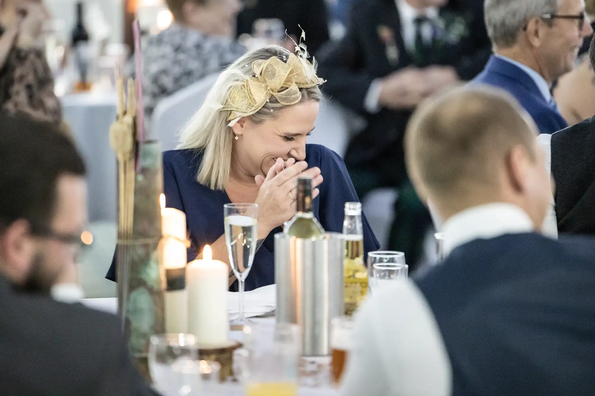 A woman in a navy outfit with a decorative fascinator laughs, covering her mouth with her hand at a formal event. She sits at a table with drinks, candles, and bottles, surrounded by other well-dressed guests in a lively, elegant setting.