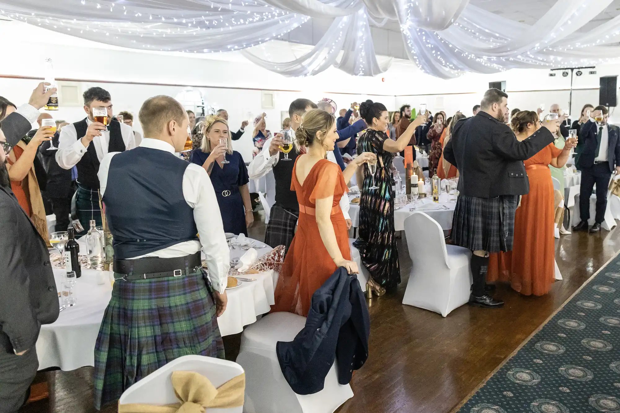 A group of people in formal attire, many in kilts and orange dresses, stand around decorated tables at a wedding reception, raising glasses in a toast under draped white fabric and fairy lights.