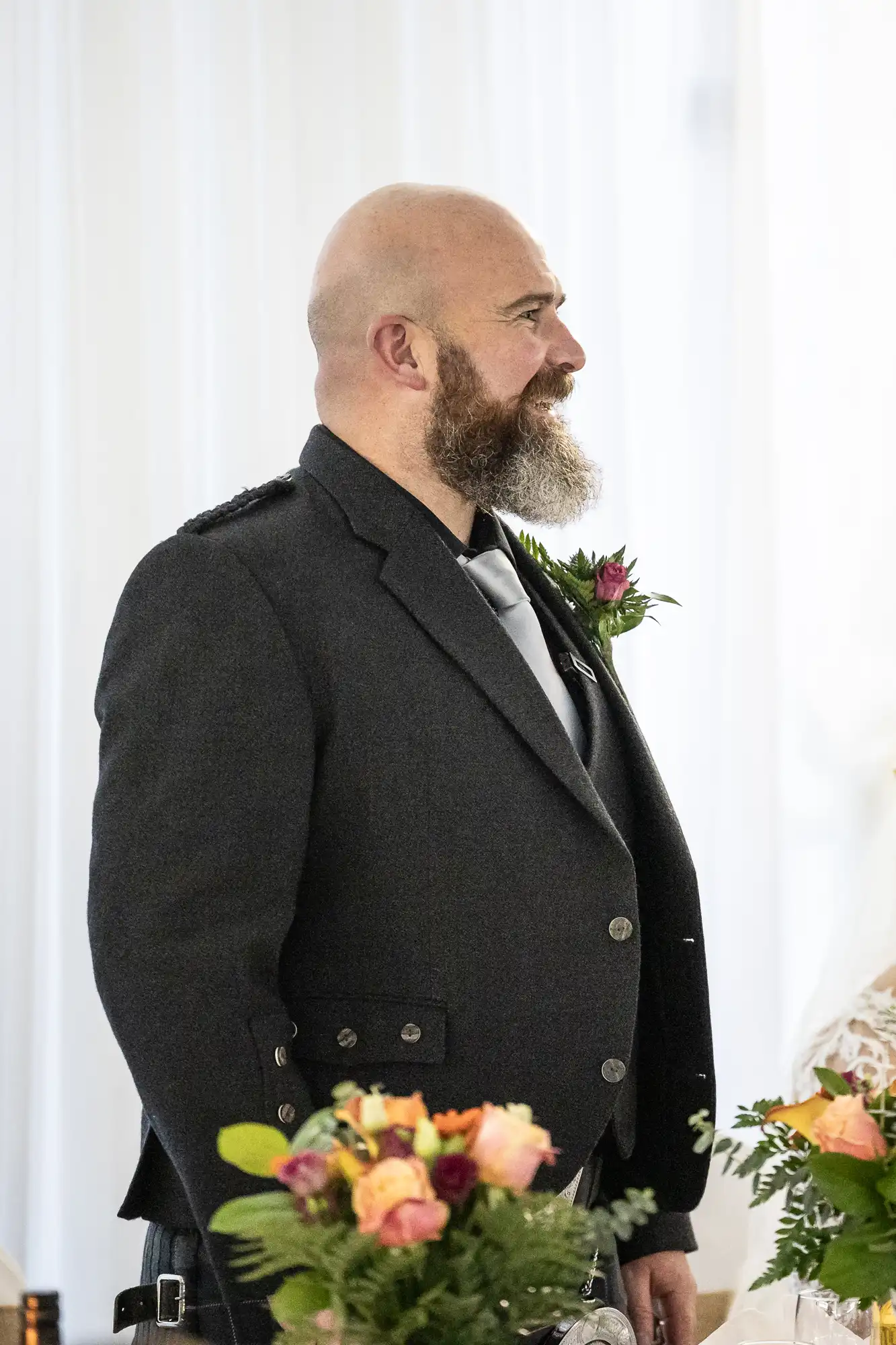 A bearded man in a dark suit with a buttonhole stands indoors beside a bouquet of orange and yellow flowers. He looks to the right, smiling slightly, against a bright, softly lit background with white curtains.