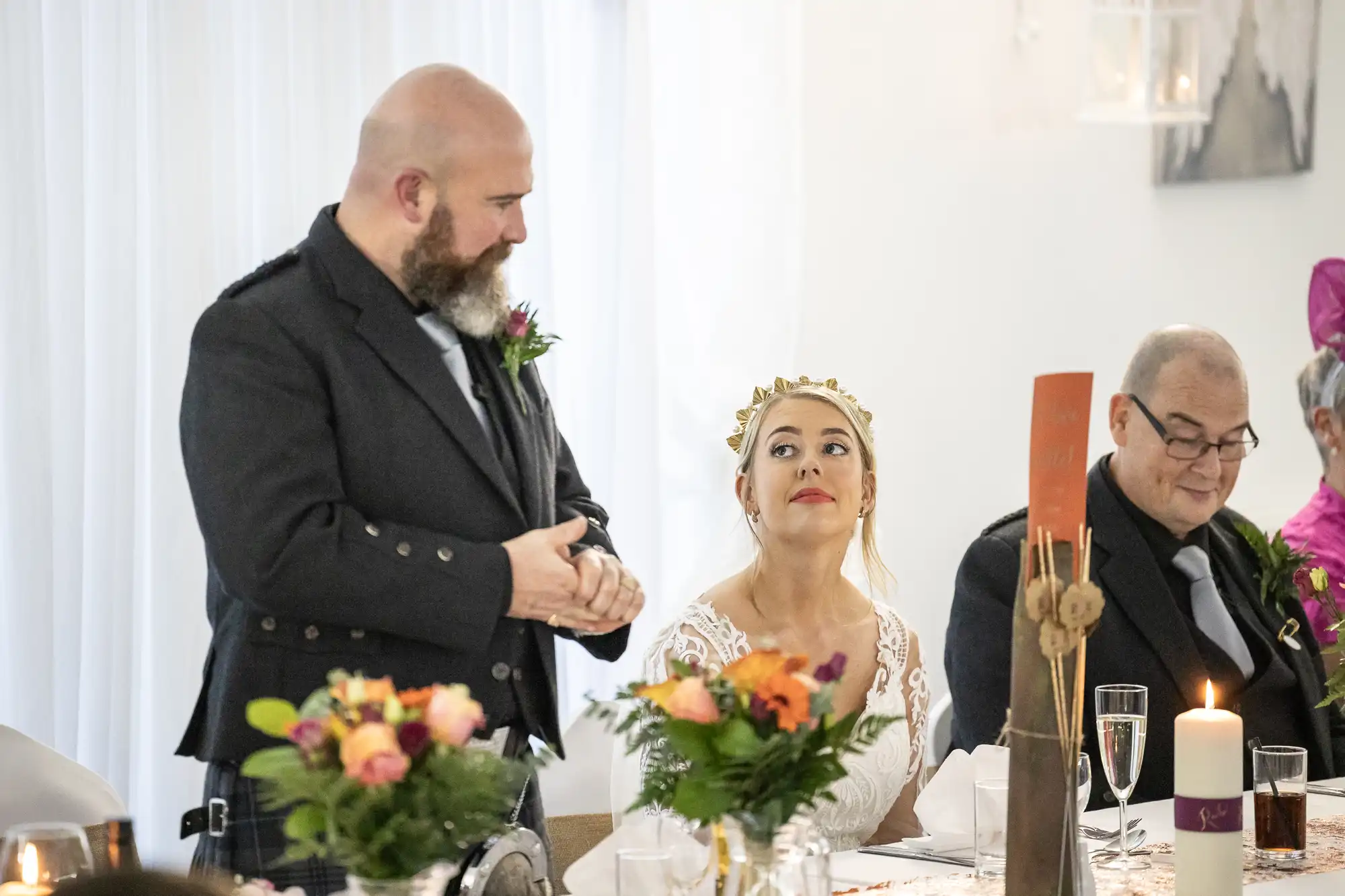 A bearded man in a dark jacket stands speaking at a table, while a bride in a white dress and gold crown looks up at him. An older man in a suit sits beside her. The table is set with flowers, candles, and champagne glasses.