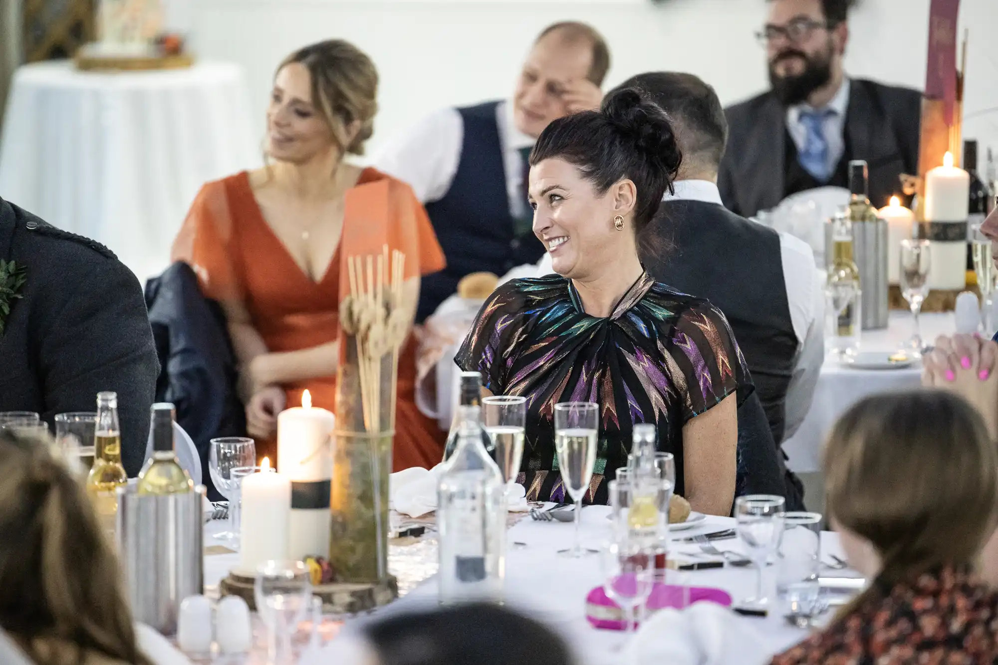 Guests dressed in formal attire sit at round tables decorated with candles, drinks, and rustic centrepieces. A woman in a colourful dress smiles, engaging with others. The atmosphere appears lively and festive, suggesting a celebration or wedding reception.