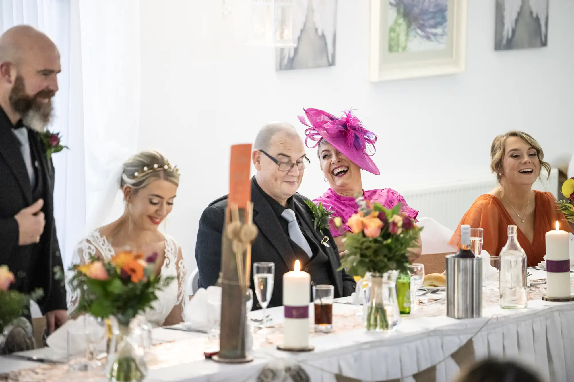 A wedding reception top table shows five people laughing and smiling. The bride in white and veil sits beside an older man in glasses and suit. A woman in bright pink and a large hat laughs, surrounded by flowers, candles, and drinks, in a bright, decorated room.