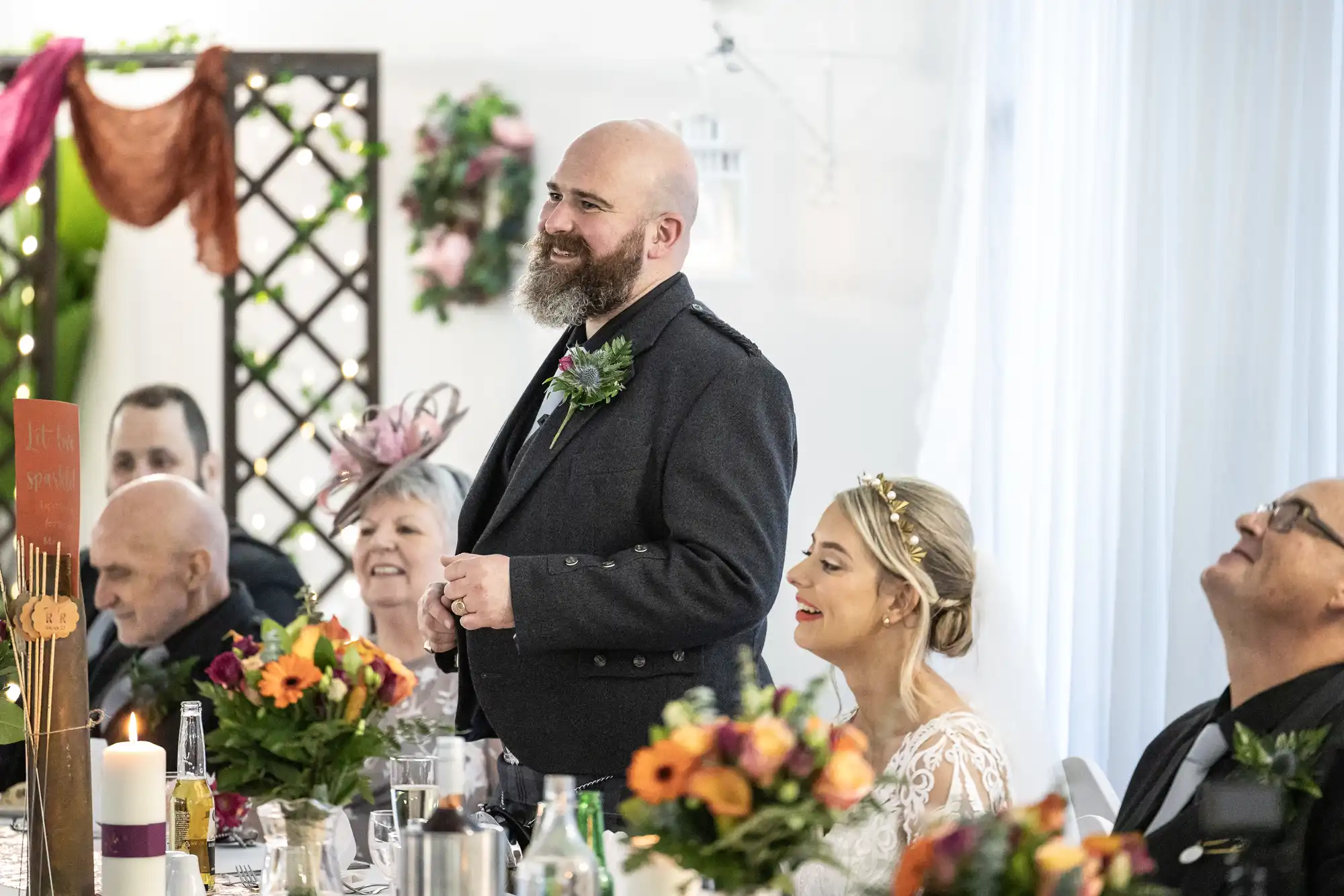 A bearded man in a dark suit stands smiling and speaking at a wedding reception table, surrounded by guests. A woman in a white lace dress, likely the bride, sits beside him, smiling. The table has floral centrepieces and candles; the atmosphere is joyful.