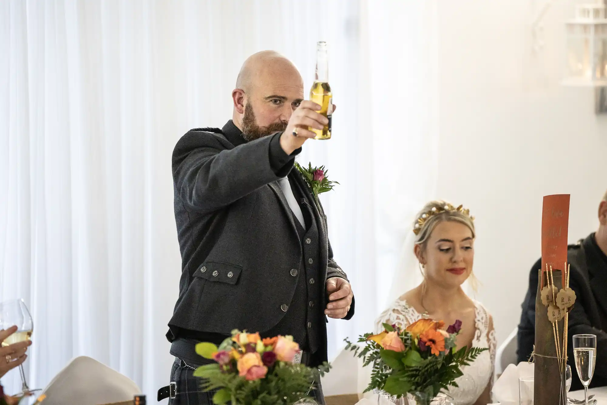 A bearded man in a dark suit raises a beer bottle in a toast at a wedding reception. Next to him, a woman in a white dress and floral headpiece sits, smiling. Colourful flower arrangements decorate the elegant, light-filled setting.
