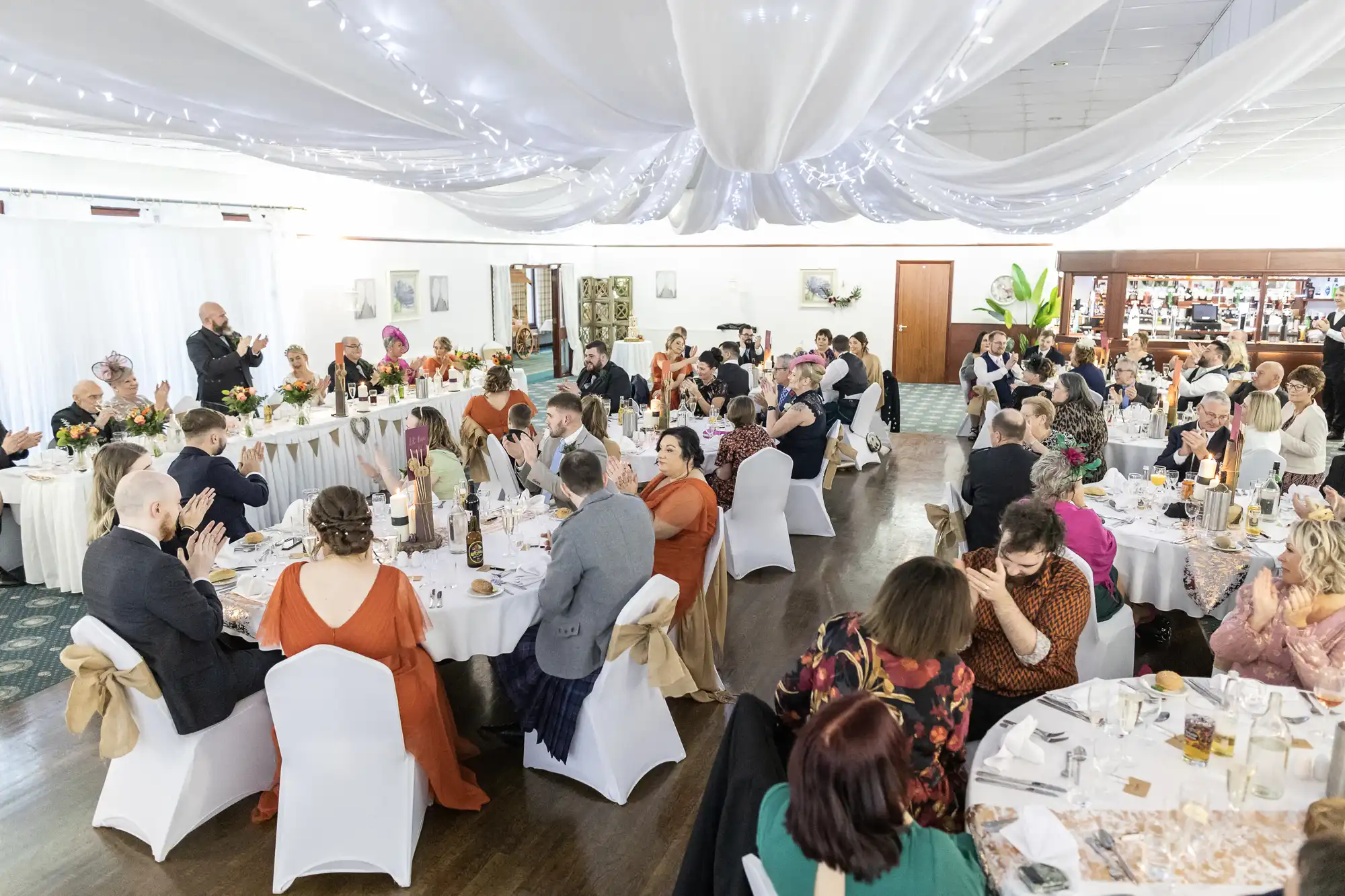 A wedding reception in a decorated hall with white draped ceilings and fairy lights. Guests in formal attire are seated around round tables, eating, talking, and applauding. The atmosphere is lively and celebratory with floral centrepieces.