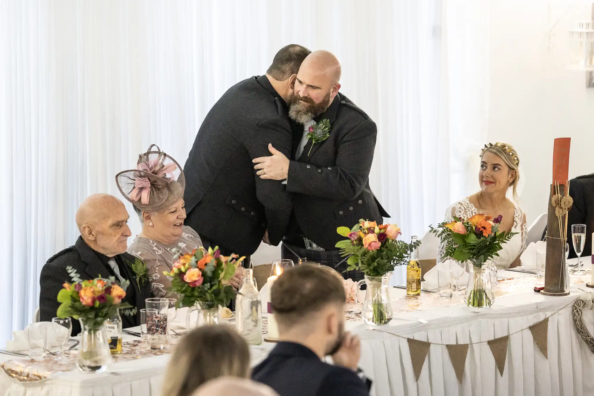 A bearded man in a dark suit stands and hugs another man at a wedding reception table decorated with flowers. Guests, including an older couple and a bride in a white dress, sit and watch, smiling. The setting is bright with white decor and crockery.