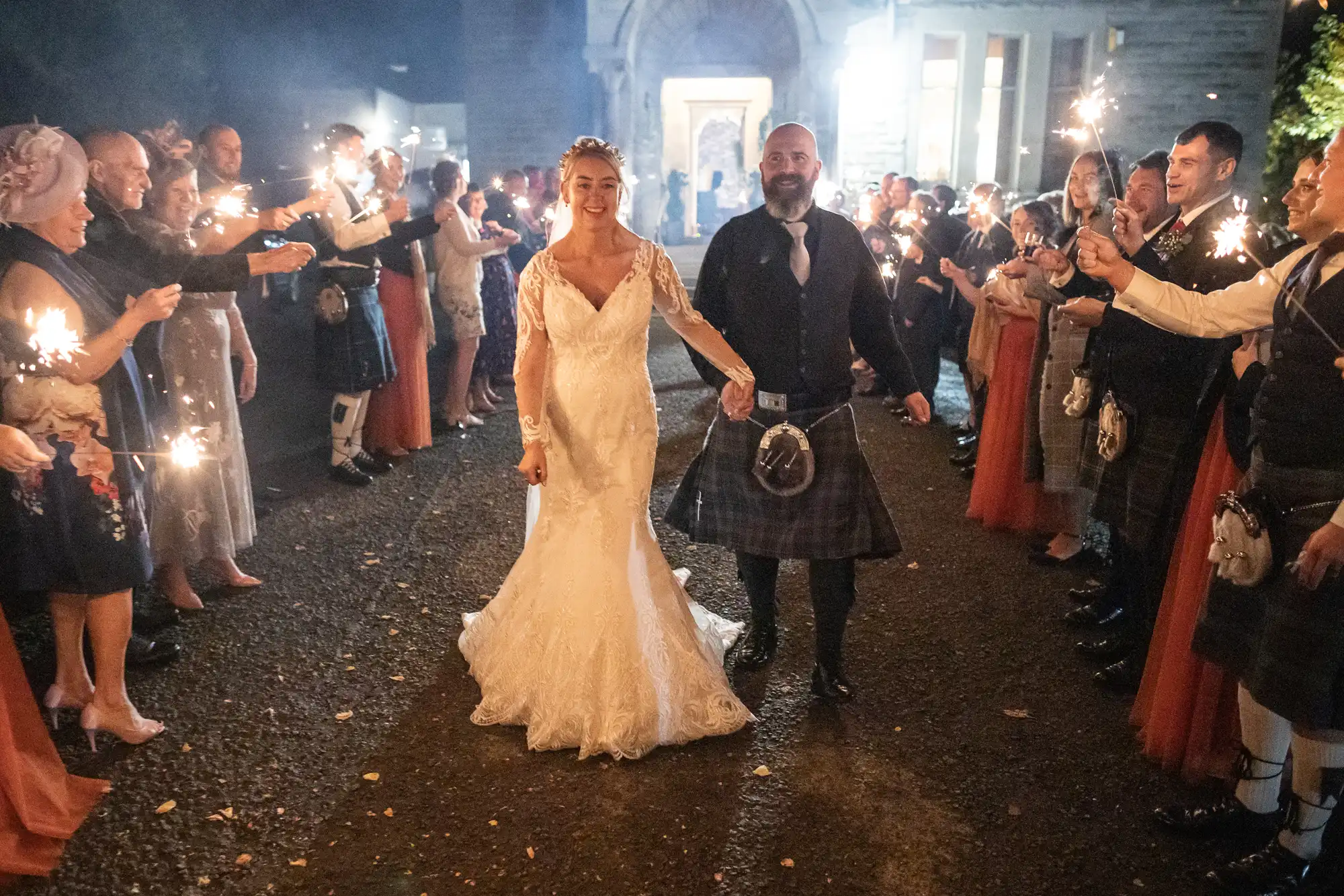 A bride in a white gown and a groom in a kilt walk hand in hand through a crowd of guests holding sparklers at night, smiling as they celebrate outside a stone building, with warm lights glowing in the background.