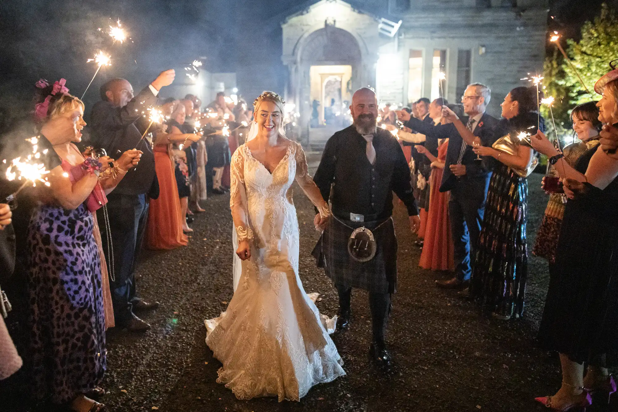 A bride in a white lace dress and a groom in a kilt hold hands, smiling as they walk outside at night between cheering guests who hold sparklers. The scene is festive, with a lit building in the background and people dressed in colourful outfits.