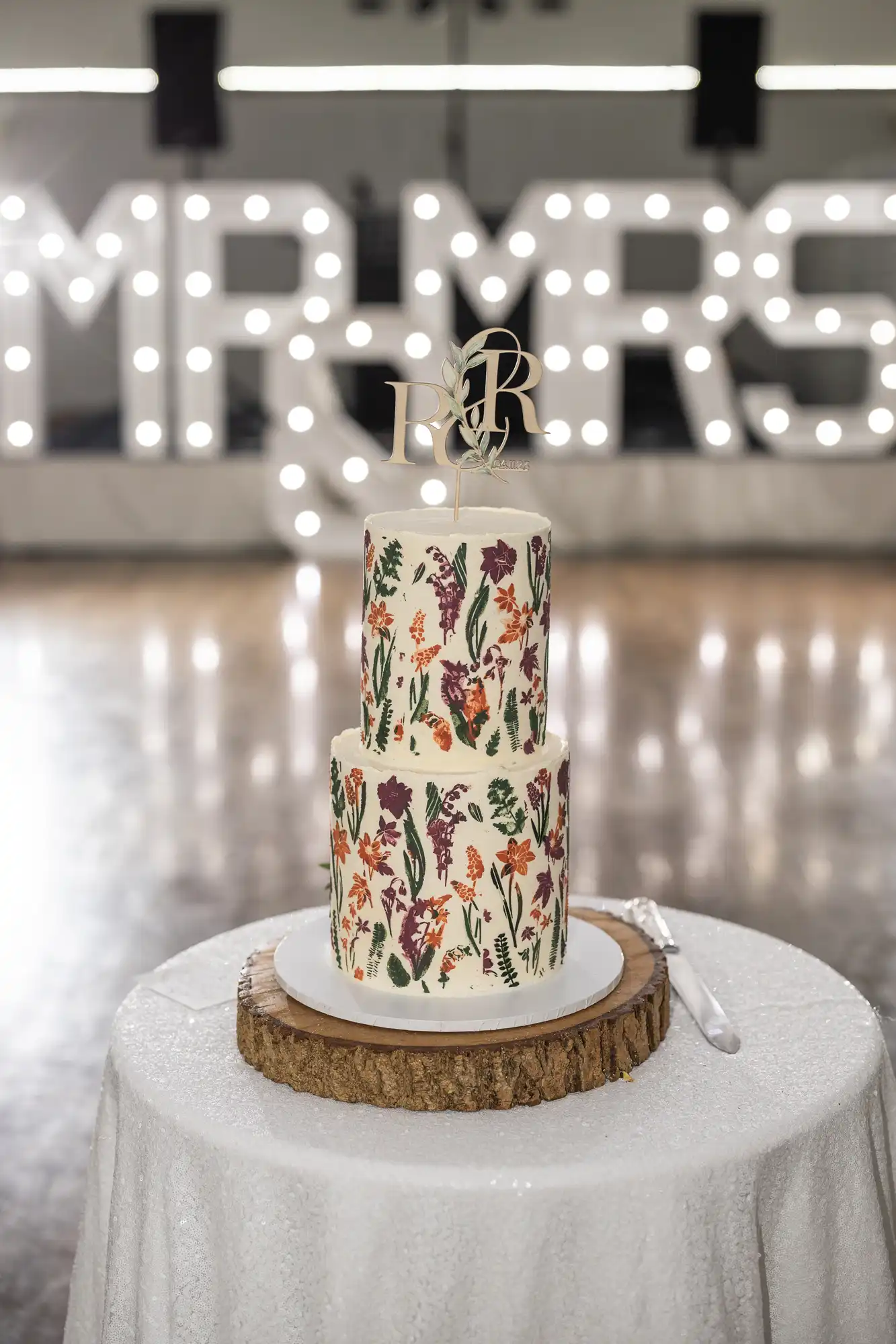 A two-tiered cake with a floral design sits on a wooden slab atop a white tablecloth. A gold monogramme cake topper is on top. In the blurred background, large marquee letters spell “MR & MRS” with bright lights. A cake knife lies beside the cake.