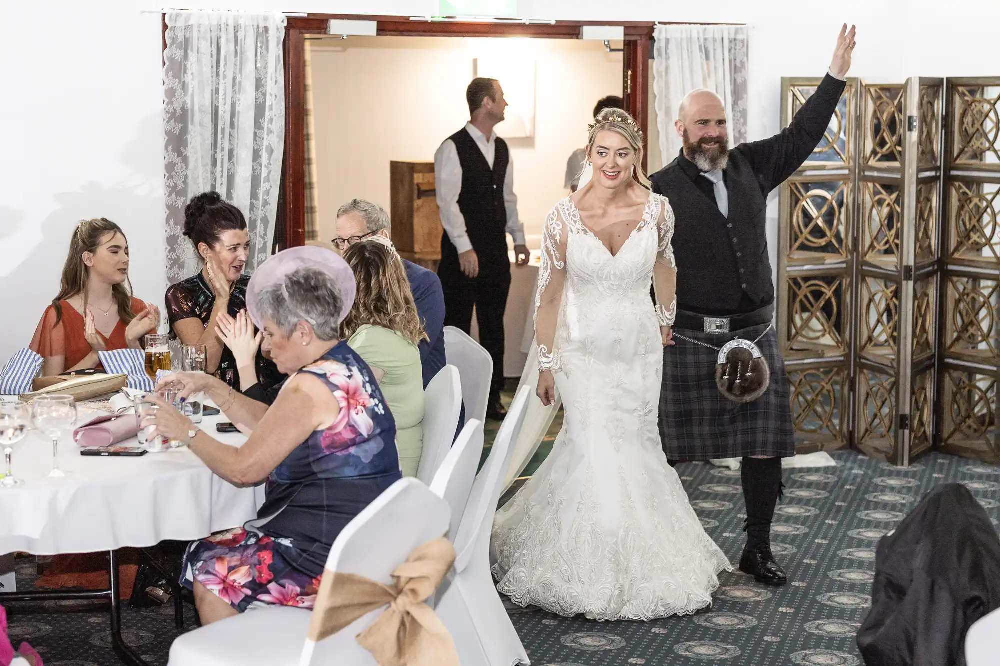 A bride in a white lace dress and a smiling man in a kilt enter a decorated reception room. Guests seated at round tables watch and react cheerfully. The atmosphere is joyful, with white chair covers and soft lighting.