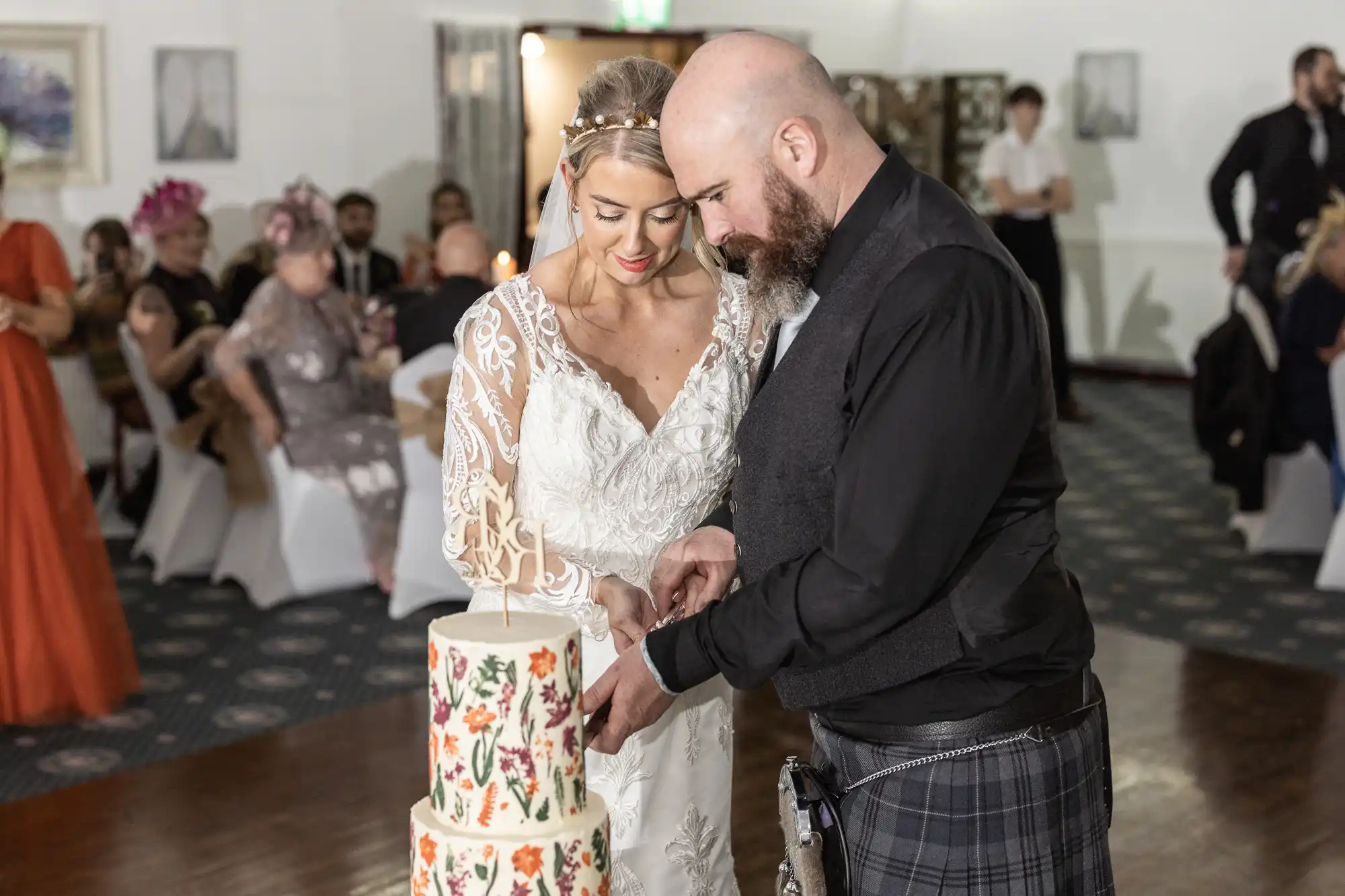 A bride in a white lace dress and a groom in a dark waistcoat and tartan kilt cut a floral wedding cake together. Guests sit and watch in the background. The setting is a formal indoor venue with white decor.