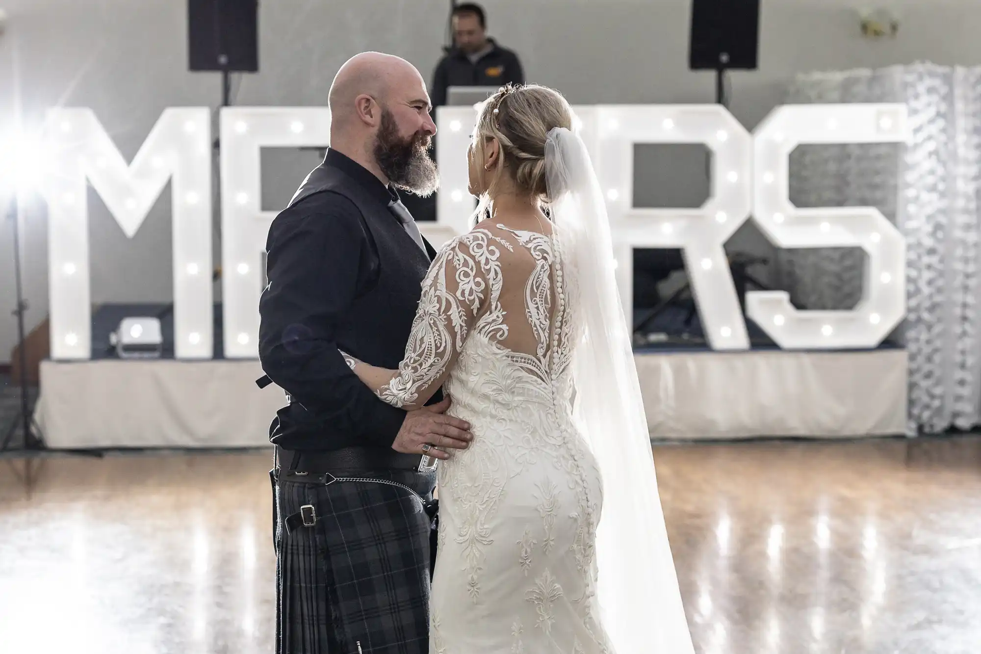 A bride in a white lace gown and veil dances with a bearded groom in a black shirt and tartan kilt. They gaze at each other while large illuminated "MR & MRS" letters glow in the background on a wooden floor.
