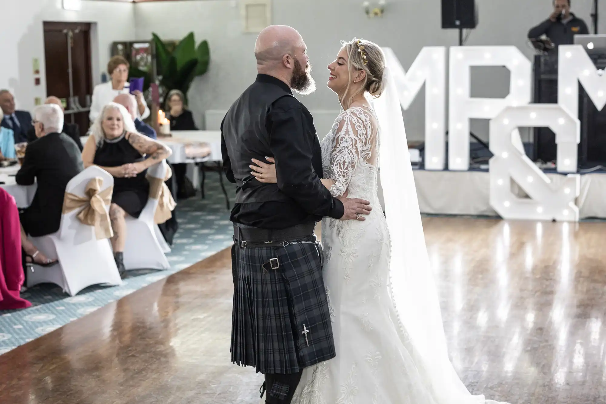 A bride in a white lace gown and veil smiles while dancing with a groom wearing a black shirt and tartan kilt. They are on a wooden dance floor, watched by seated guests. Large illuminated “MR & MRS” letters are in the background.
