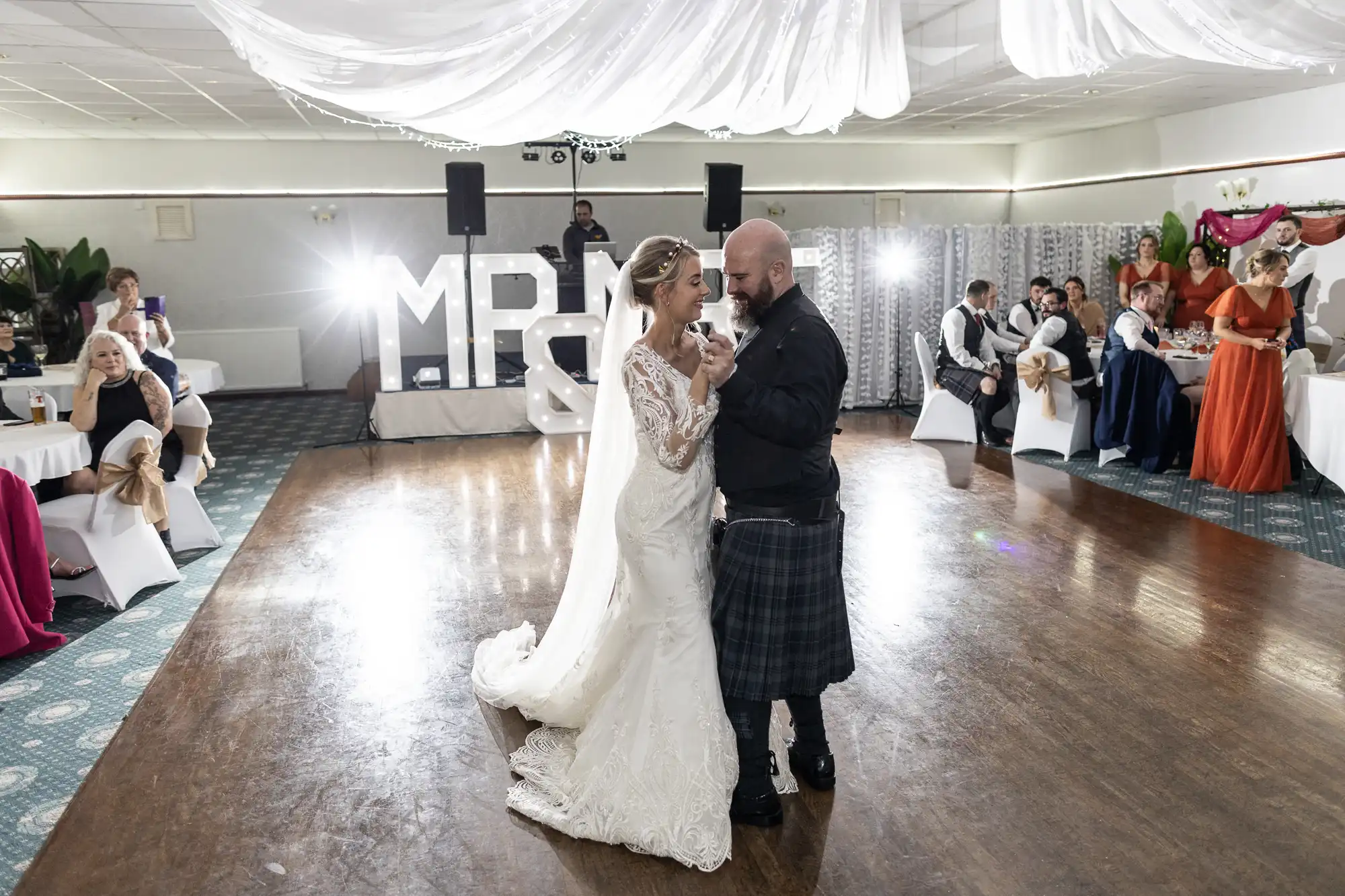 A bride in a white lace gown and a groom in a kilt share their first dance in a reception hall with guests seated around. Large illuminated “MR & MRS” letters and a DJ are in the background, with white drapery on the ceiling and bright lighting.