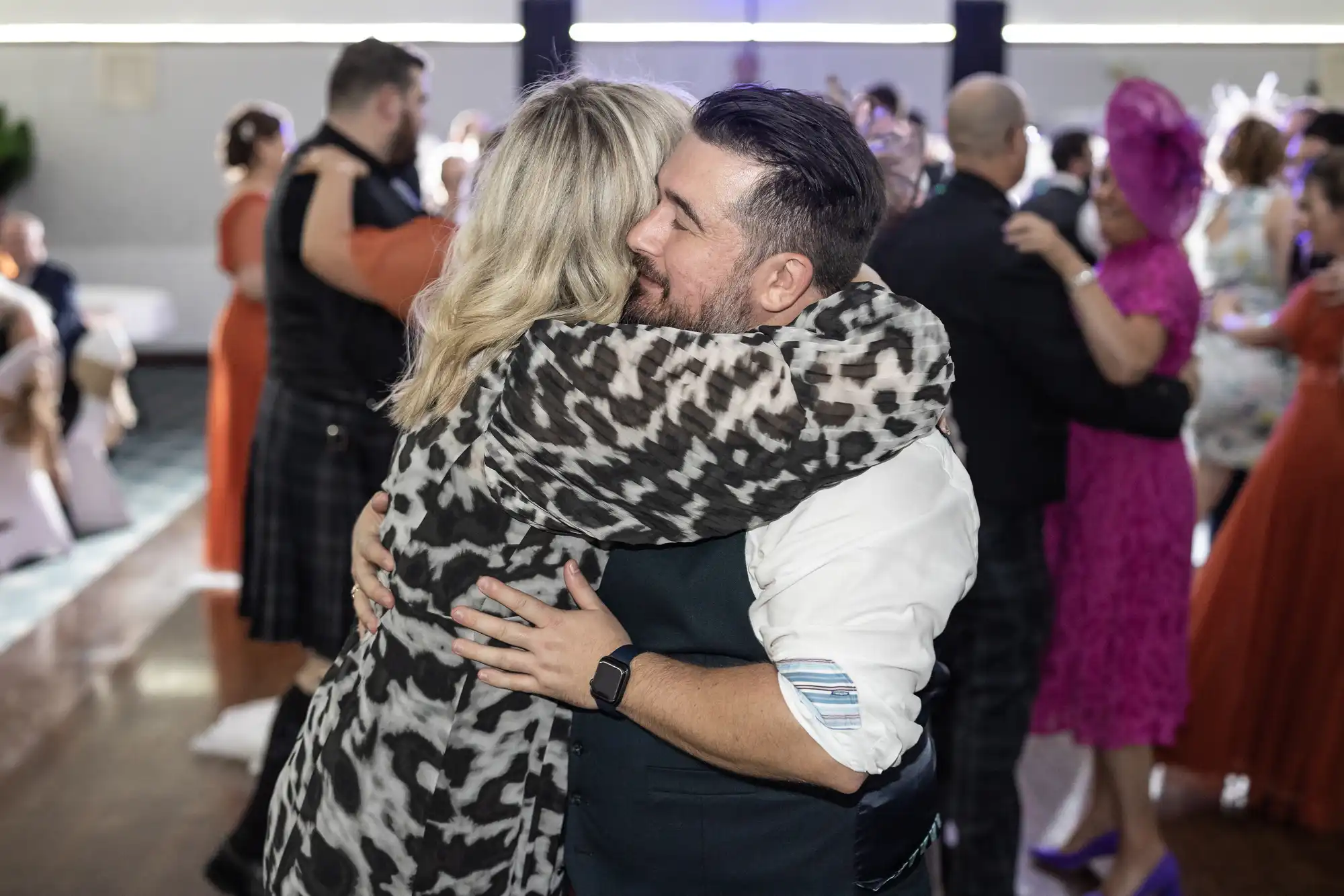 A man and a woman embrace warmly on a dance floor at a lively event. The woman has blonde hair and wears a leopard print outfit. The man has dark hair and a beard, wearing a white shirt and navy waistcoat. Other couples dance and mingle in the background.