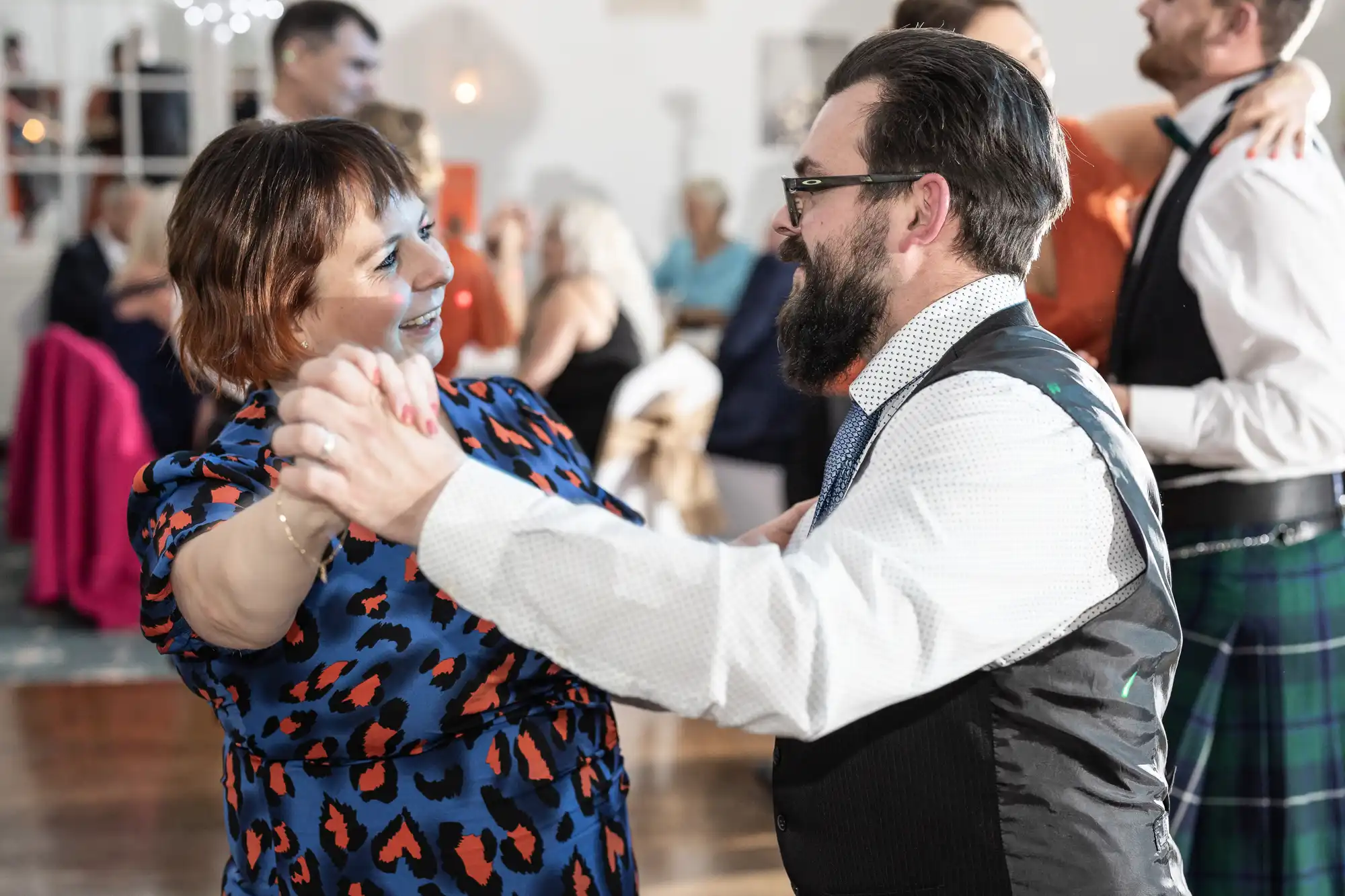 A woman in a blue and orange dress and a bearded man in glasses and a waistcoat smile warmly at each other while dancing. They hold hands, mid-step, at a lively, well-lit event with other people dancing in the blurred background.