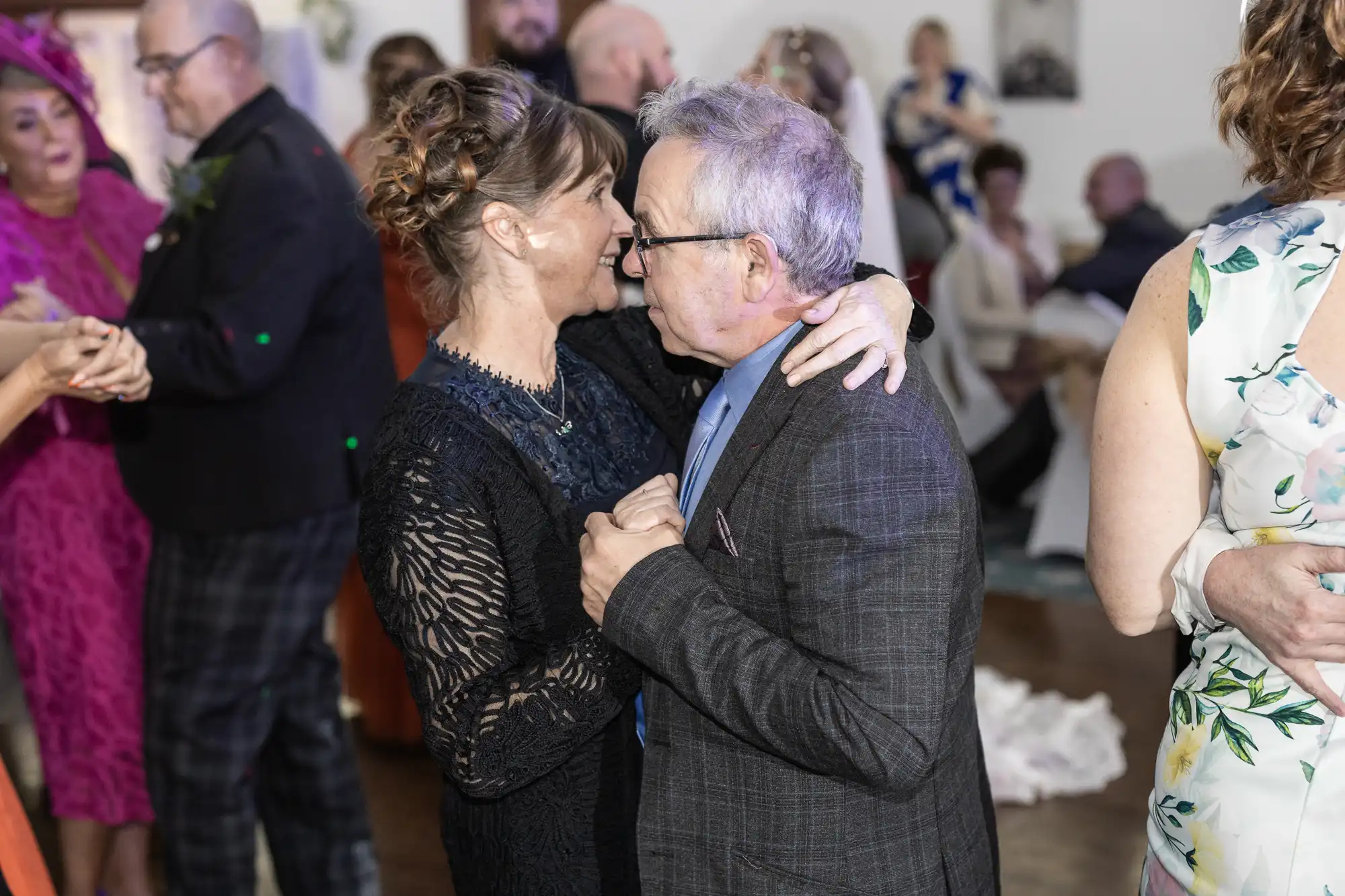 An older couple in formal attire lovingly dance close together at a wedding reception, smiling and gazing at each other. Other couples dance in the background, and guests in colourful dresses and suits fill the softly lit room.