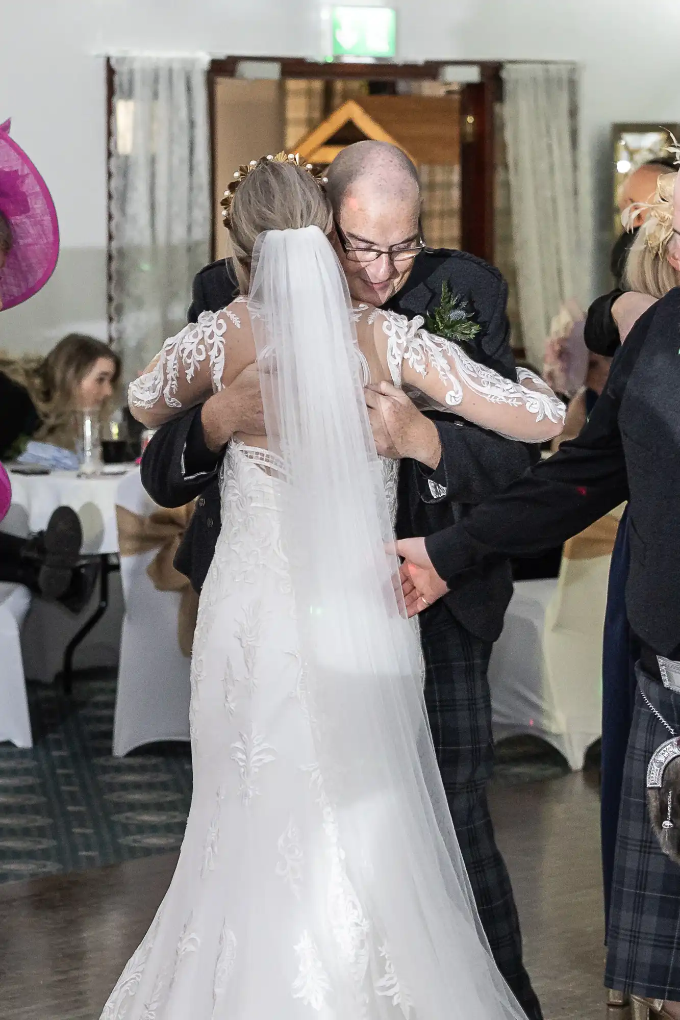 A bride in a white lace gown and veil hugs an older man in a suit at an indoor wedding reception. Guests sit at decorated tables in the background. The embrace appears warm and emotional, with both central figures at the focus of the scene.