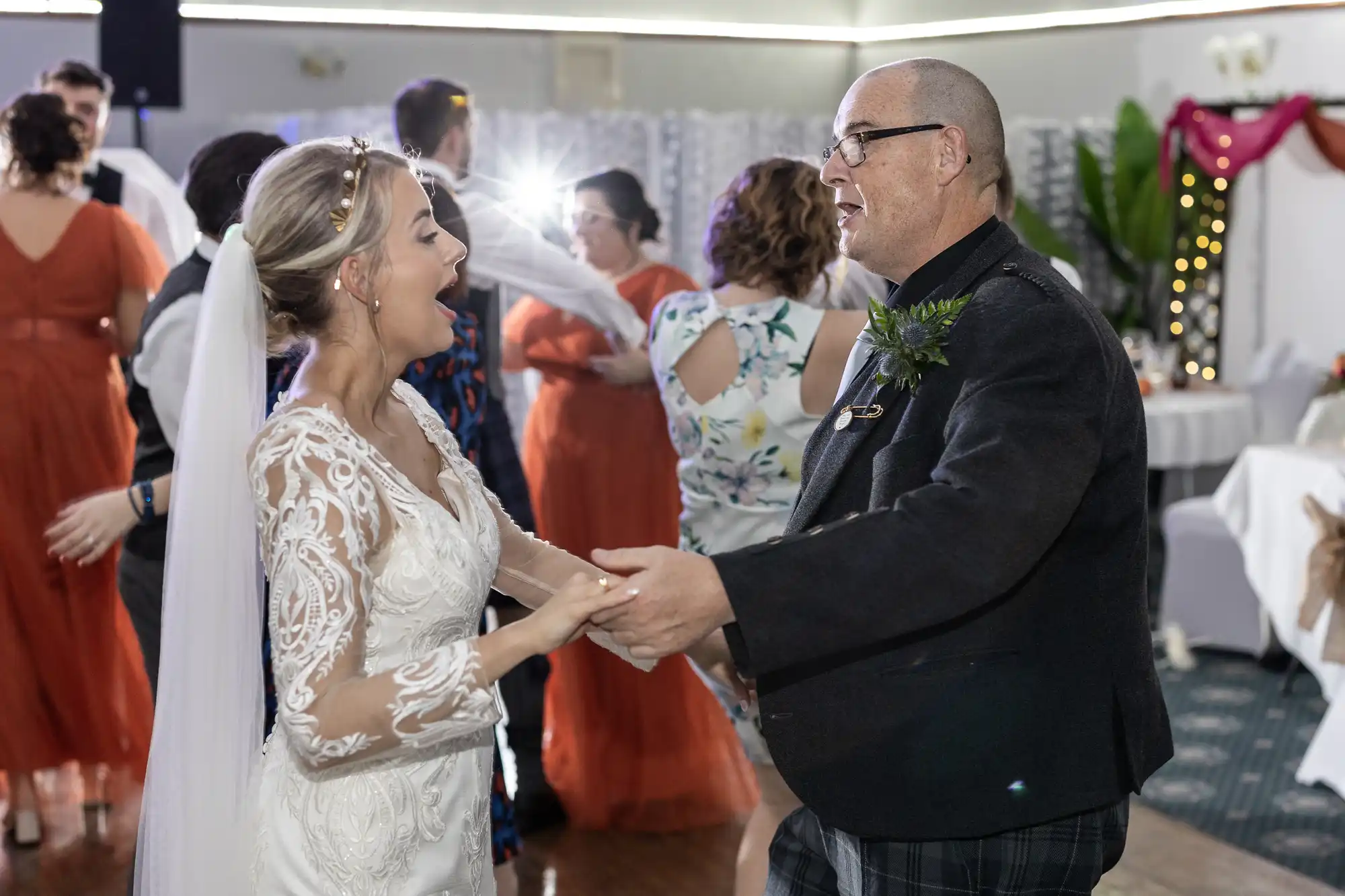 A bride in a white lace dress and veil dances, smiling and surprised, with an older man in glasses and a dark suit. People in colourful dresses dance in the background under bright indoor lighting.