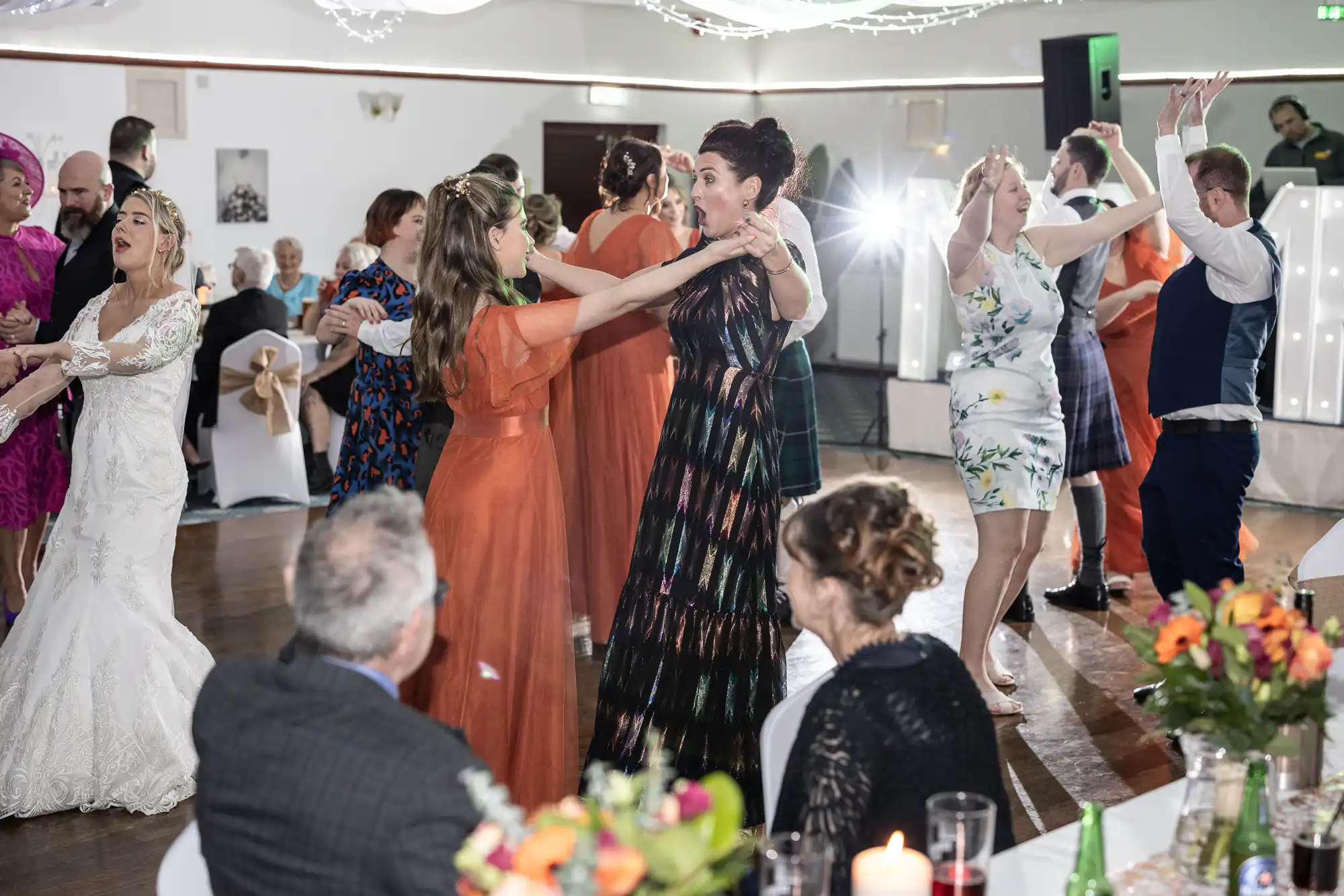 Guests dance energetically in a decorated hall with fairy lights above. Women in colourful dresses and a bride in white are in the centre, smiling and holding hands. Seated guests in the foreground watch, with flower arrangements and candles on the tables.