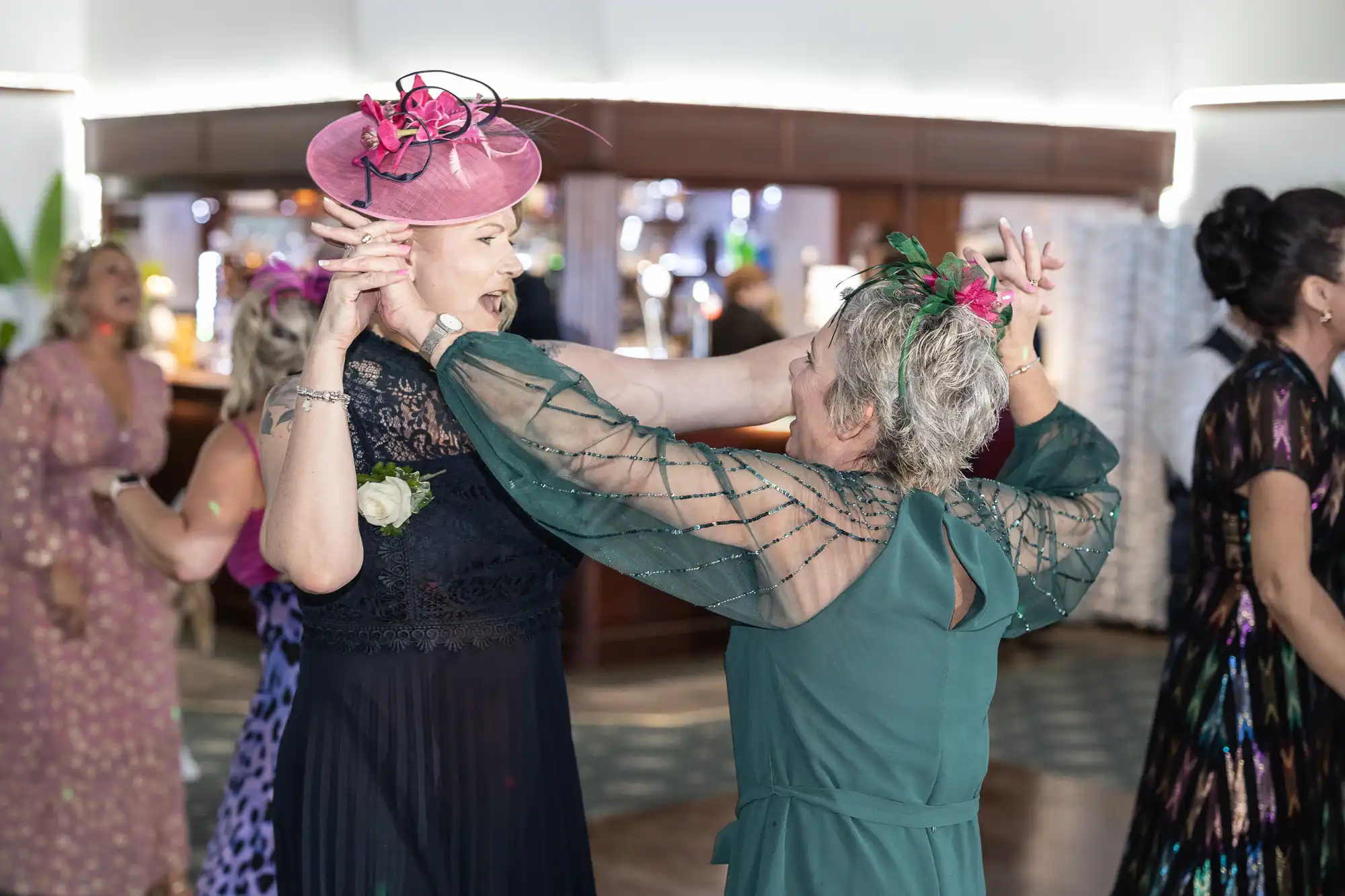 Two women in elegant dresses and colourful hats joyfully dance together at an indoor event. One wears a black dress with a pink hat, the other has a green dress with a green fascinator. Other guests in festive attire mingle in the softly lit background.