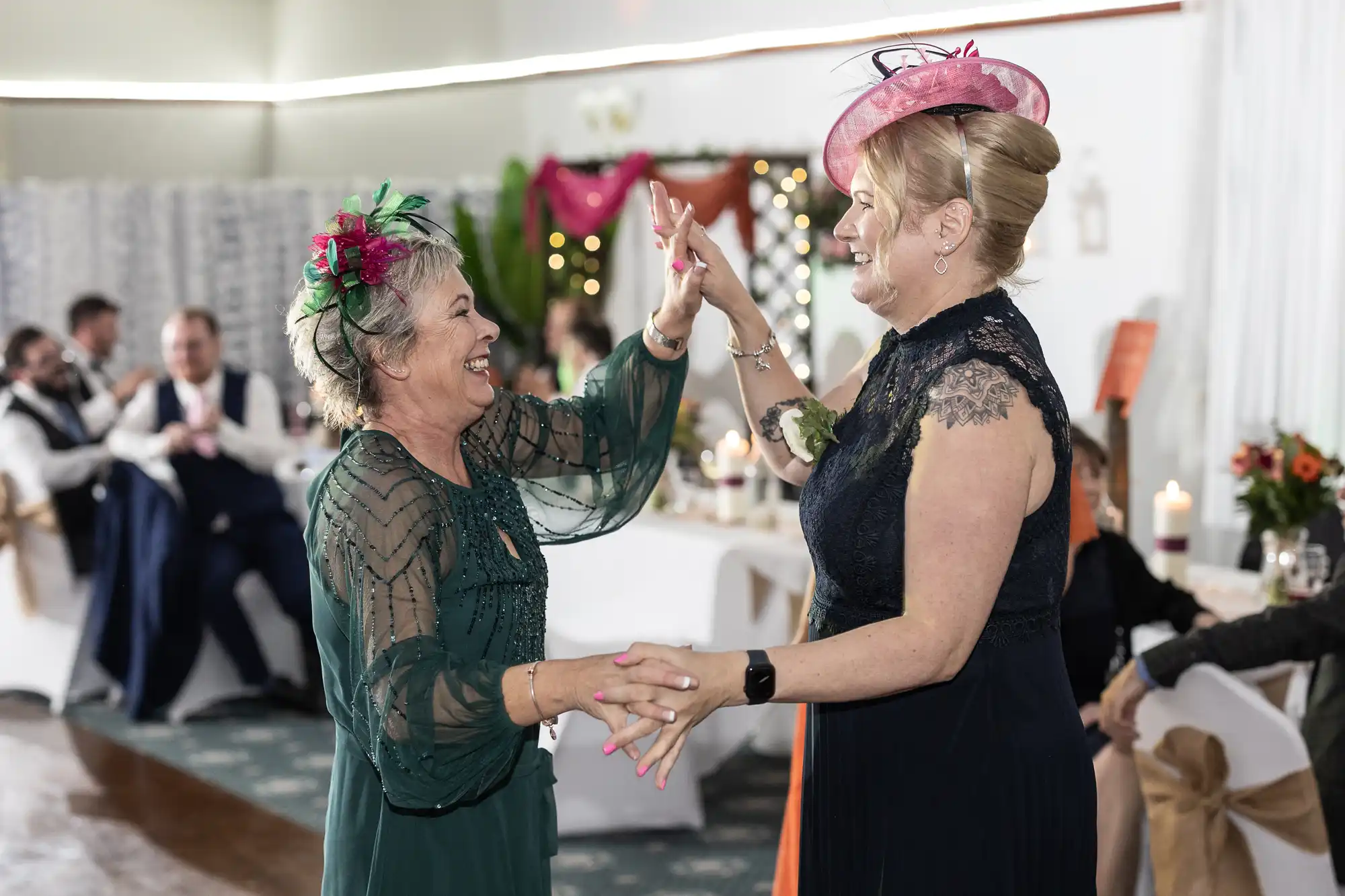 Two women in elegant dresses and floral hats are smiling and high-fiving while holding hands, dancing at a festive indoor event. Guests sit at decorated tables in the background with candles, flowers, and fairy lights. The mood is joyful and celebratory.