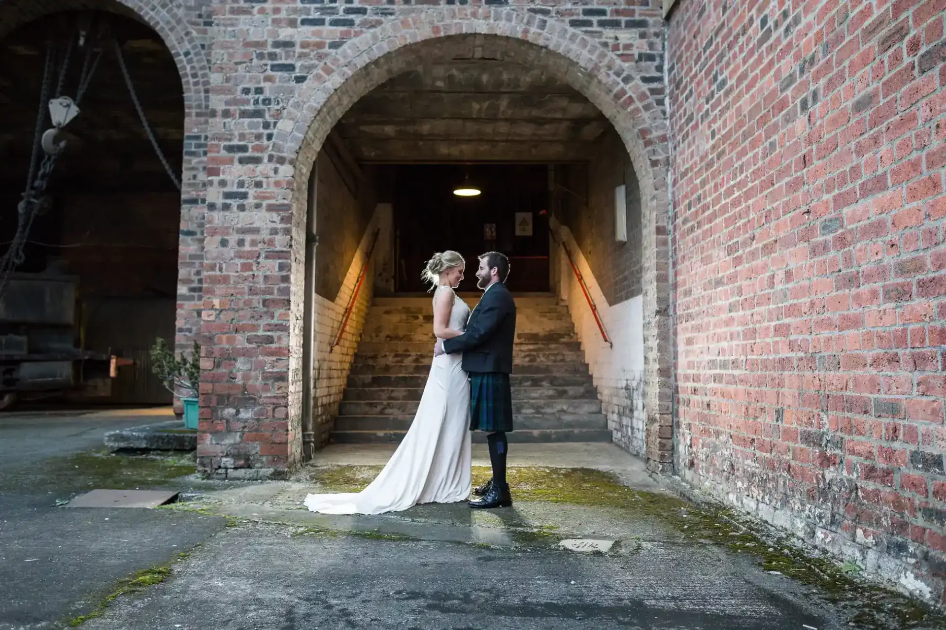 A bride in a white dress and a groom in a black jacket with a kilt stand facing each other, holding hands, under a large brick archway at National Mining Museum Scotland. Stairs with a glowing light lead upwards behind them, creating a soft, intimate atmosphere.