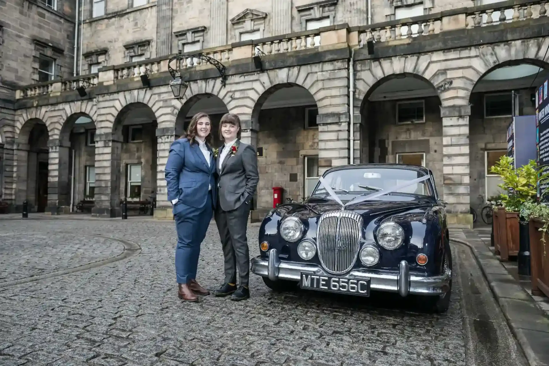 Two female brides in suits stand beside a classic dark blue car parked on a cobblestone street in front of a historic stone building at Edinburgh City Chambers.