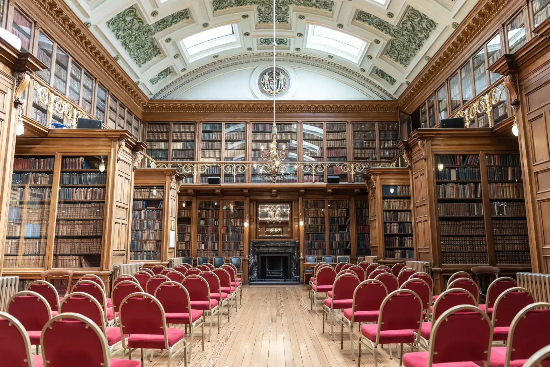 Ornate library with tall bookshelves, a decorative ceiling, a chandelier, and rows of empty red chairs facing a fireplace and lectern at Royal College of Physicians of Edinburgh.