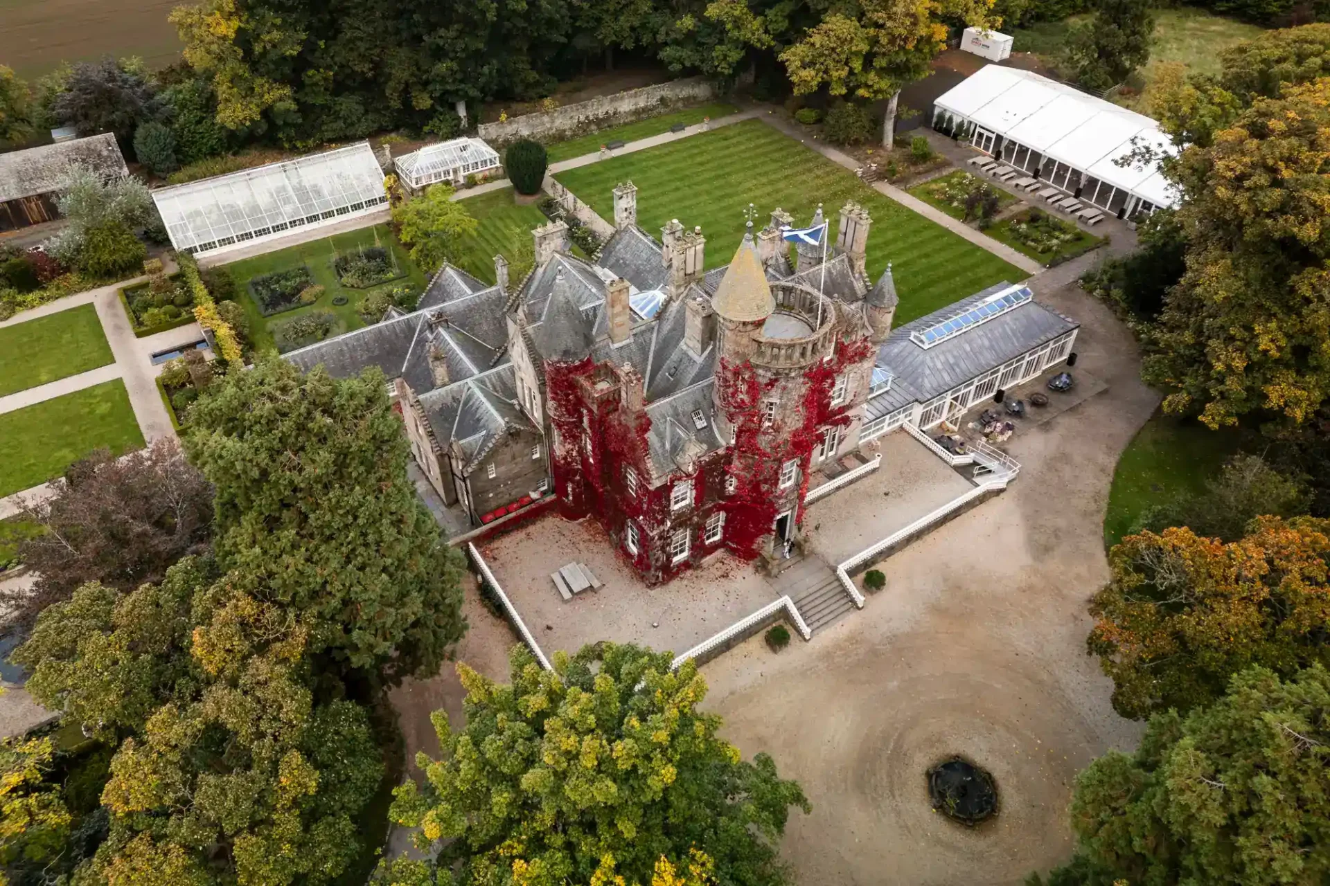 Aerial view of a historic stone mansion with red ivy on its façade, surrounded by gardens, trees, and a courtyard with a circular drive at Carlowrie Castle, Edinburgh.