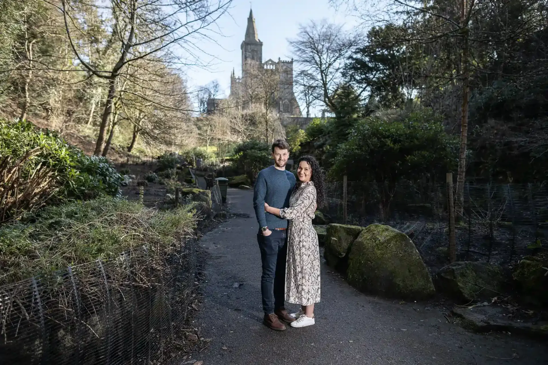 A couple stands embracing on a path in a wooded Pittencrieff Park with Dunfermline Abbey in the background. The man wears a blue jumper and dark trousers; the woman wears a floral dress and white shoes. Trees and bushes surround them, and a large stone church towers in the background under a clear sky.