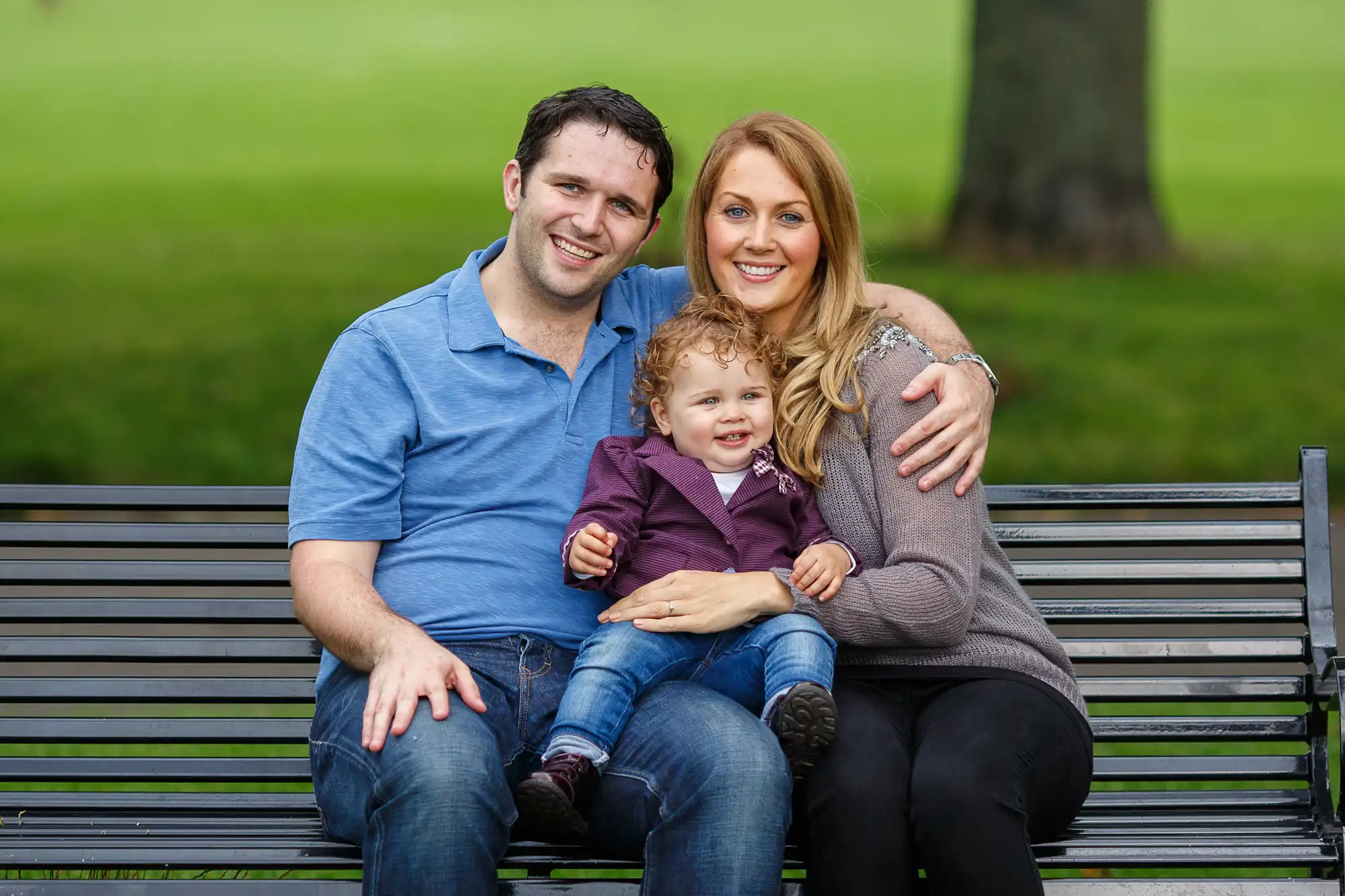 A happy family of three, with a man, a woman, and a young child, smiling and sitting closely together on a park bench.