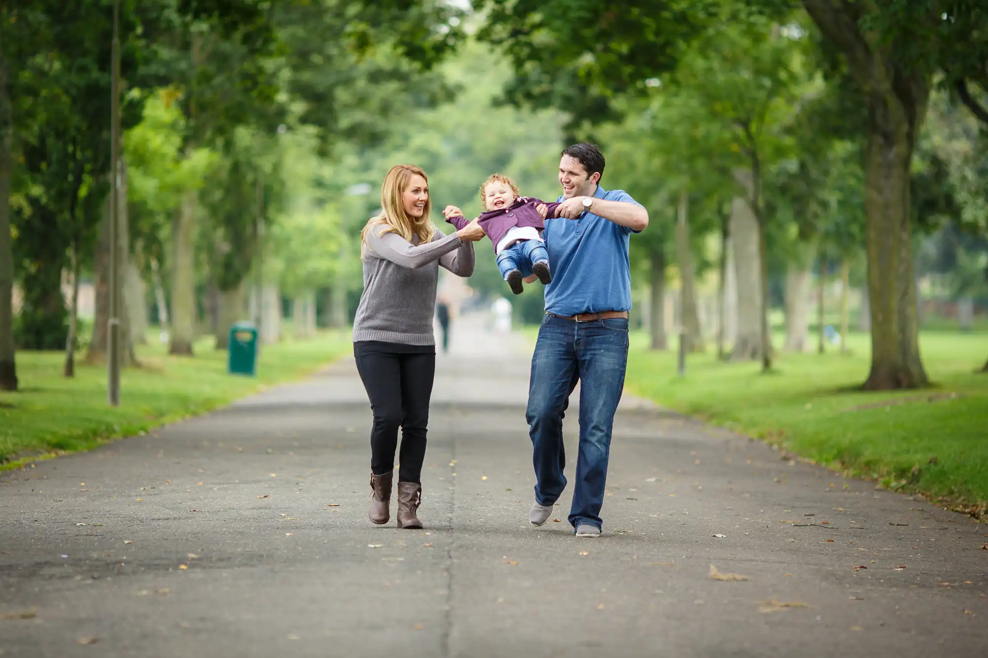 A family with a young child joyfully playing, as they walk down a tree-lined park path.